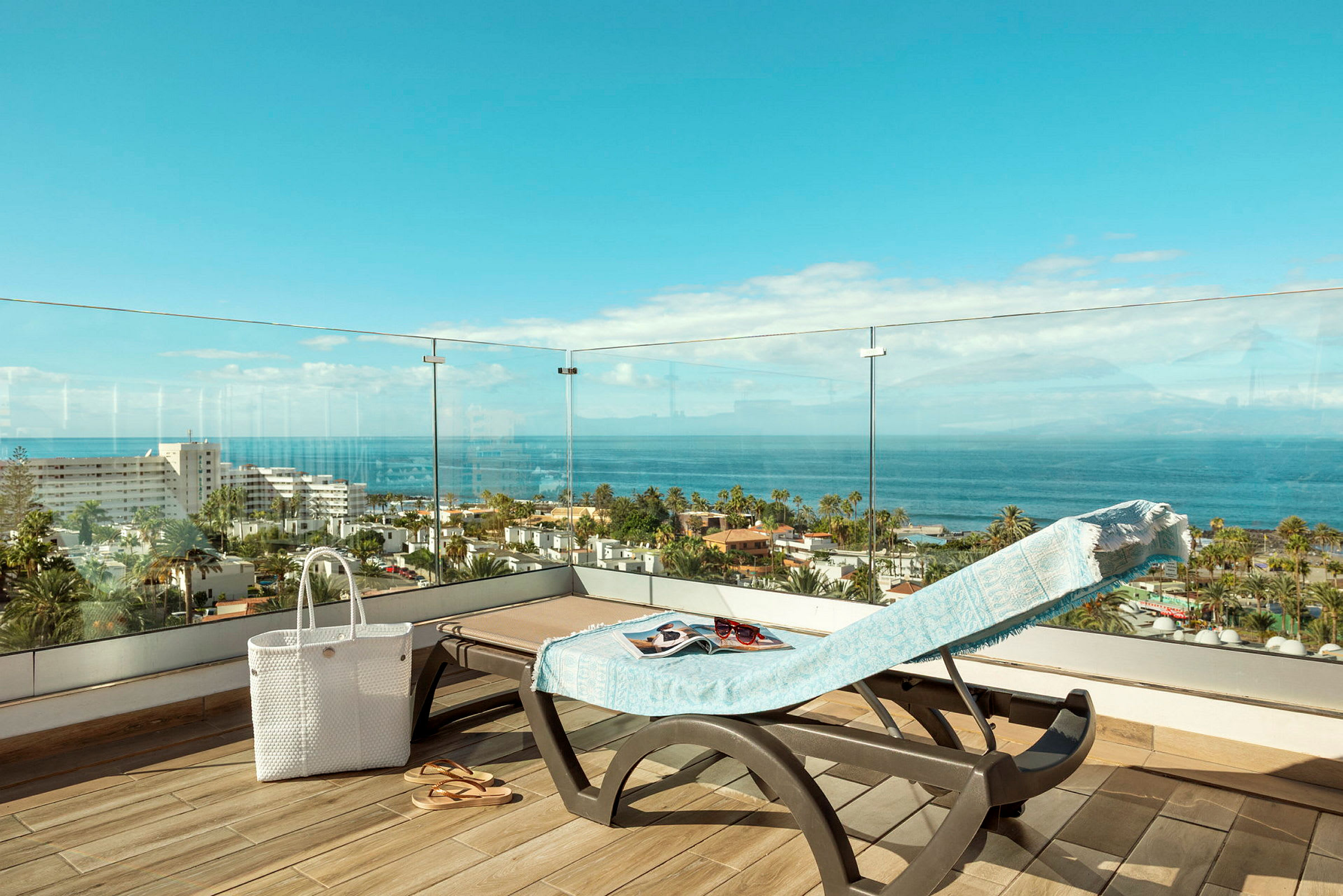 Udsigt fra en terrasse med en solseng dækket med et håndklæde, en hvid taske og sandaler, over man ser havet og bygninger. (AI-genereret tekst)