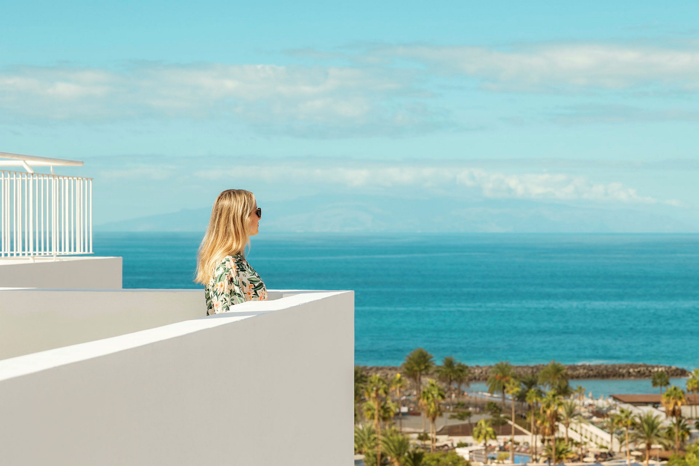 Udsigt over havet fra en høj balkon med en kvinde, der kigger mod horisonten, omgivet af en klar himmel og vegetation. (AI-genereret tekst)