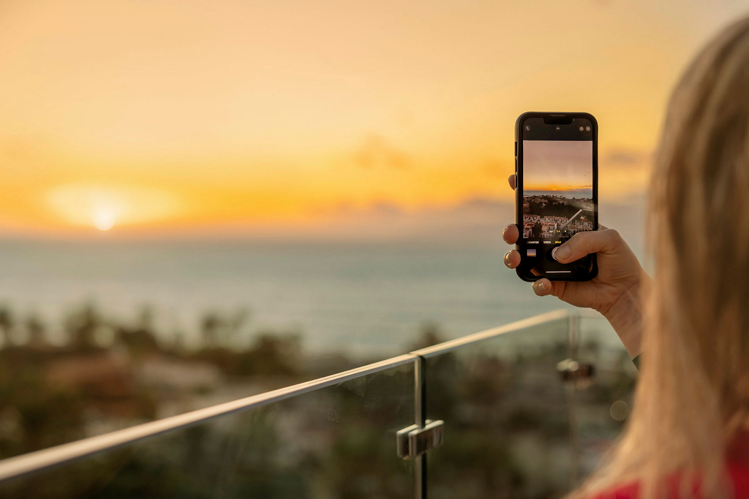 Person tager et billede med sin mobiltelefon af en solnedgang over havet fra en altan. (AI-genereret tekst)
