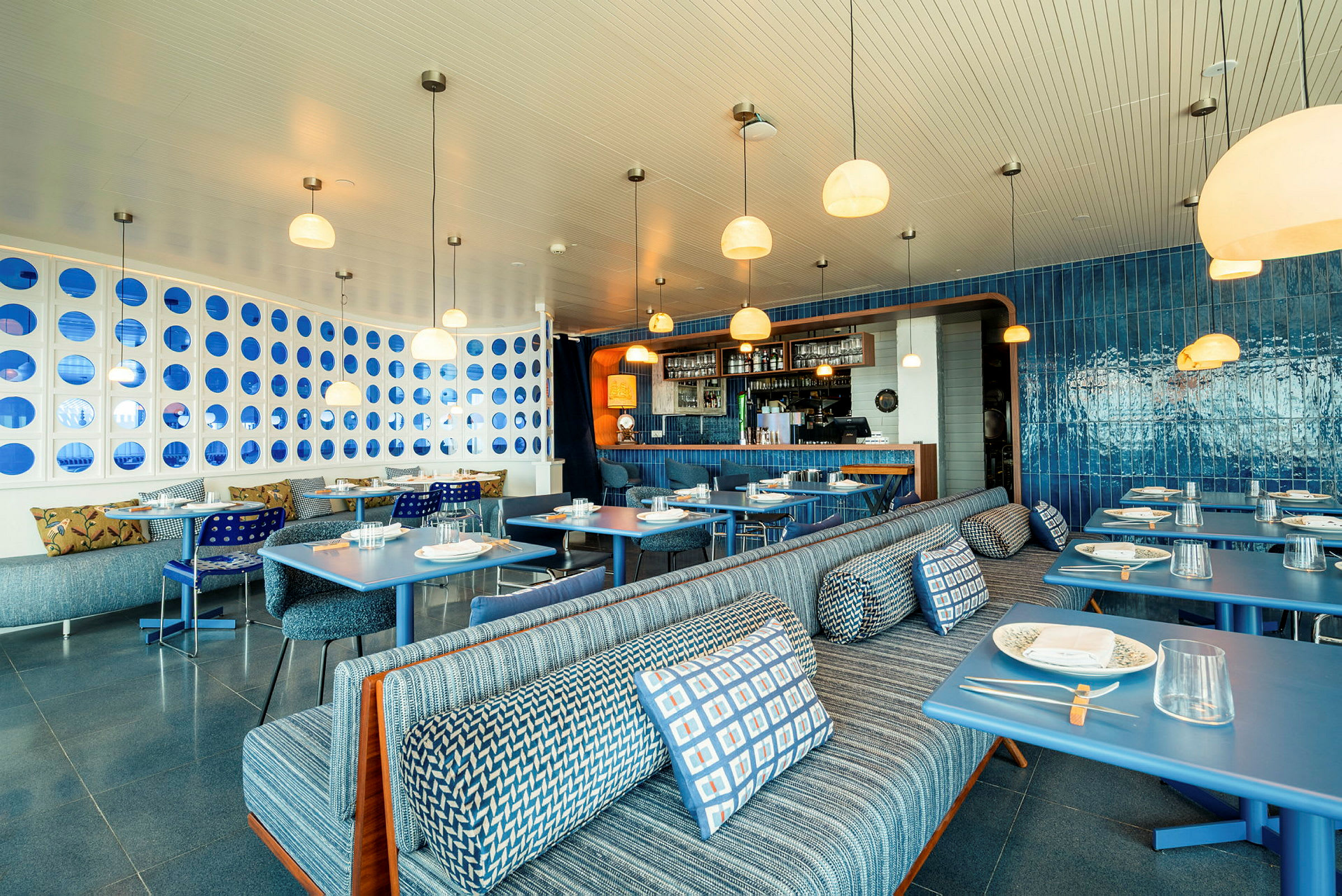 Billede af en moderne restaurant med elegant interiør i blå nuancer, siddepladser med mønstrede puder og opdækkede borde. (AI-genereret tekst)