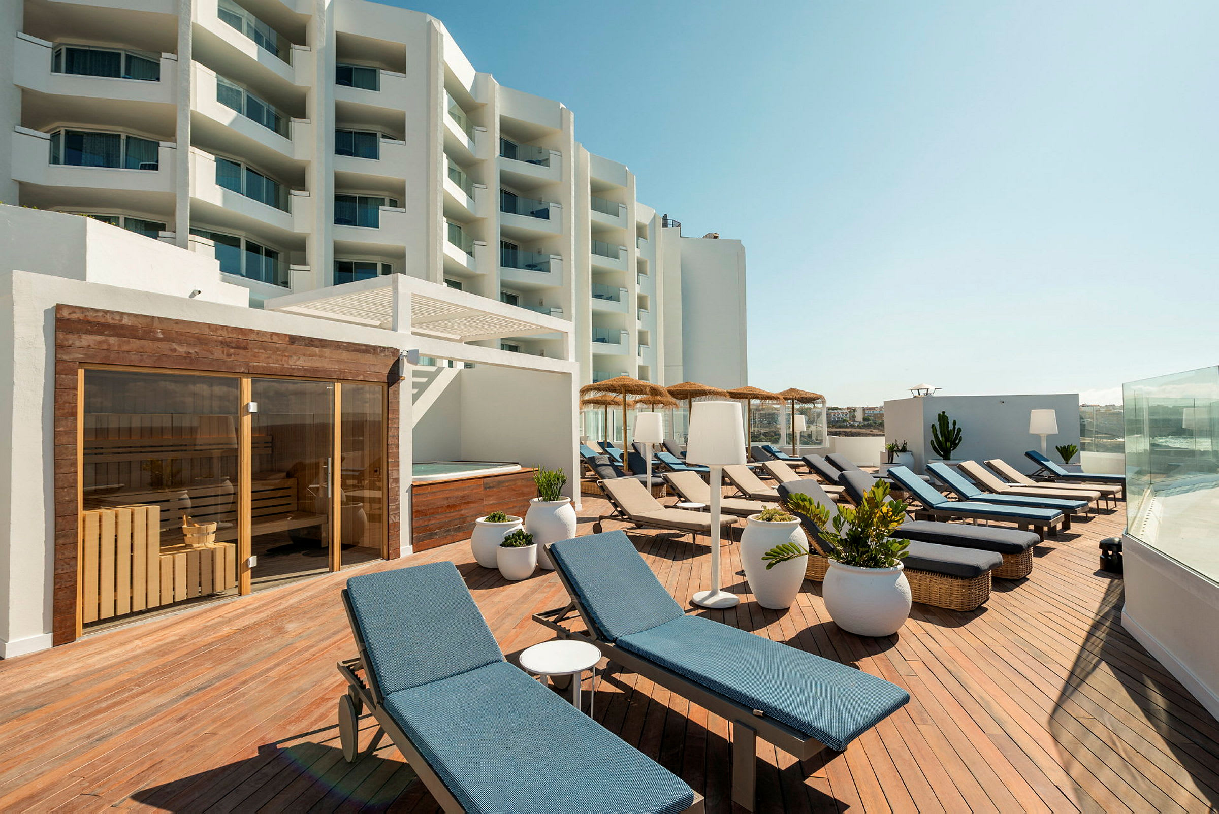 En terrasse med liggestole, parasoller og planter. I baggrunden ses et lysefarvet hotel. (AI-genereret tekst)