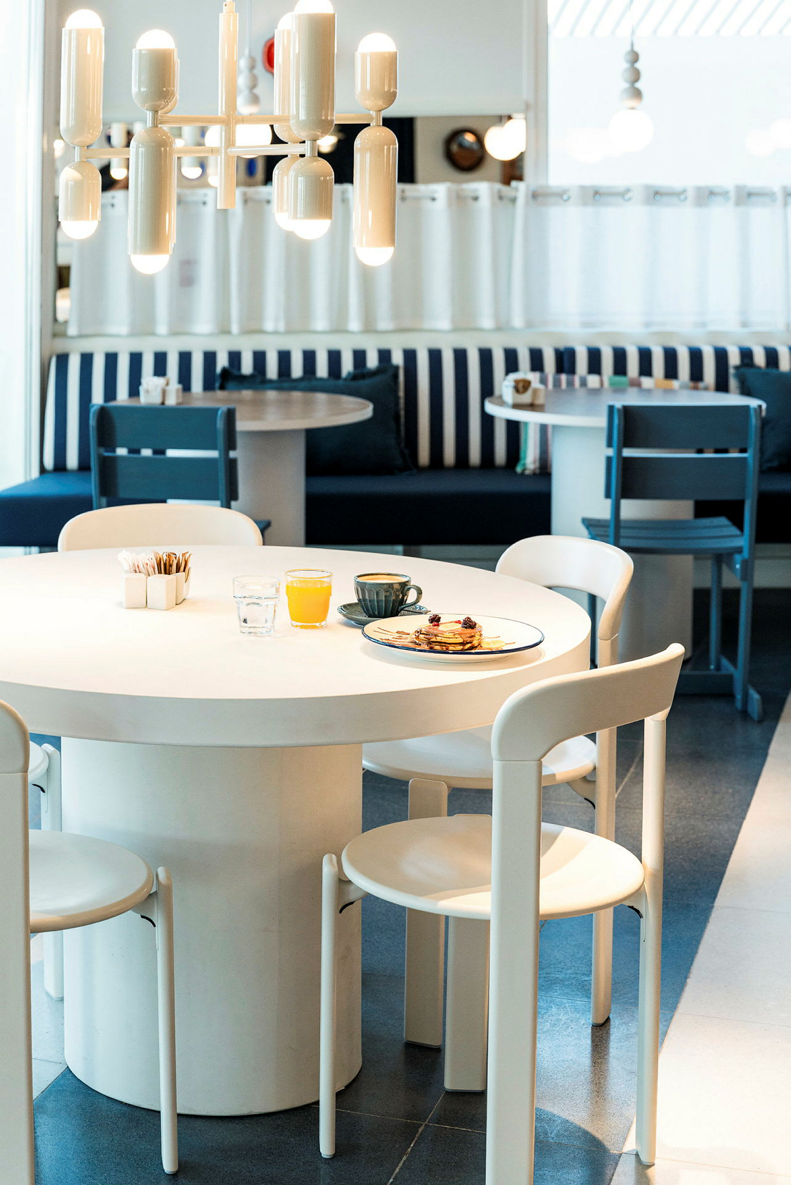 En café med hvide borde og stole, og en tallerken med pandekager samt drikkevarer står på et af bordene. (AI-genereret tekst)