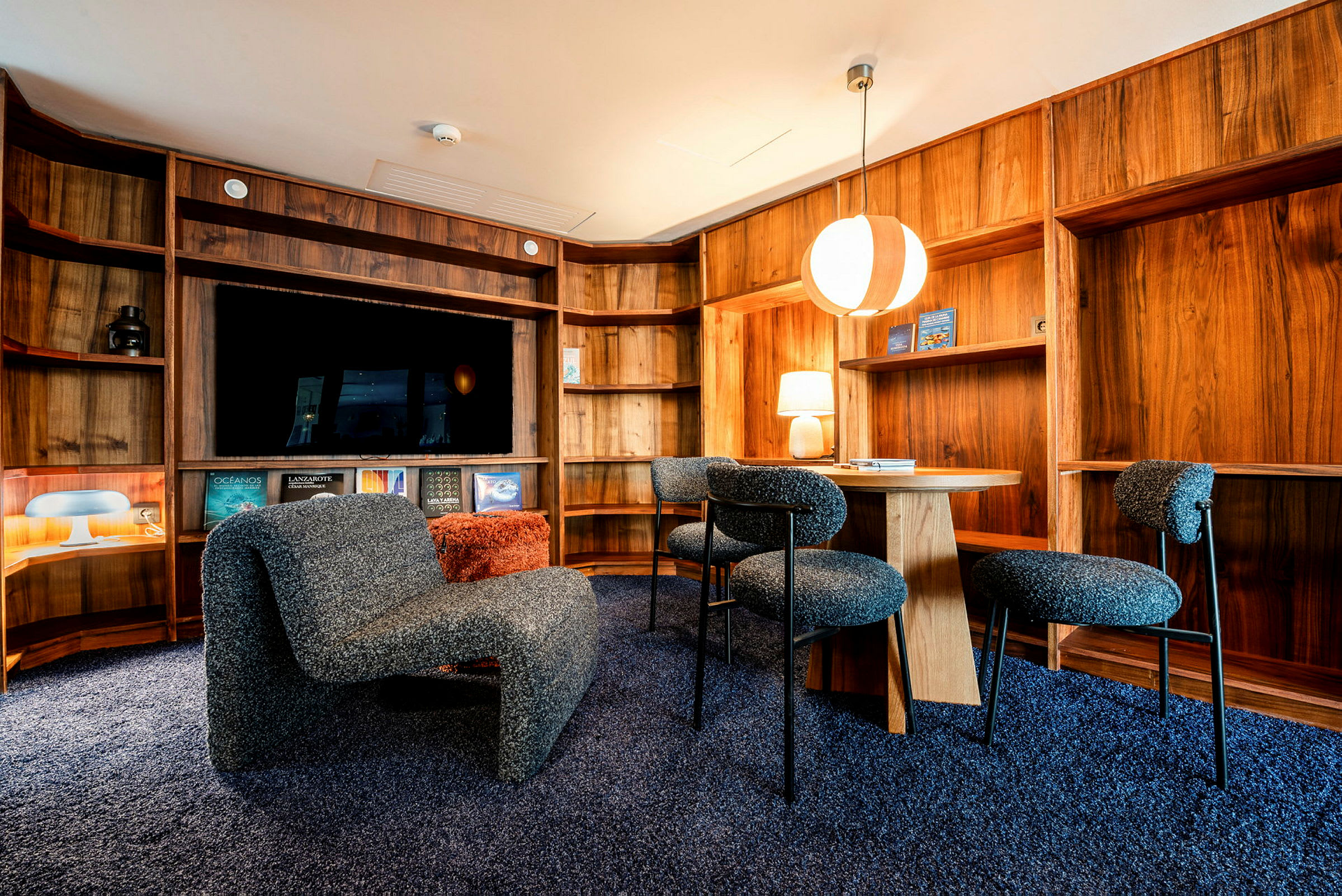 En moderne lounge med træpaneler, hylder, en siddegruppe med grå stole og bord samt en stor loftslampe. (AI-genereret tekst)
