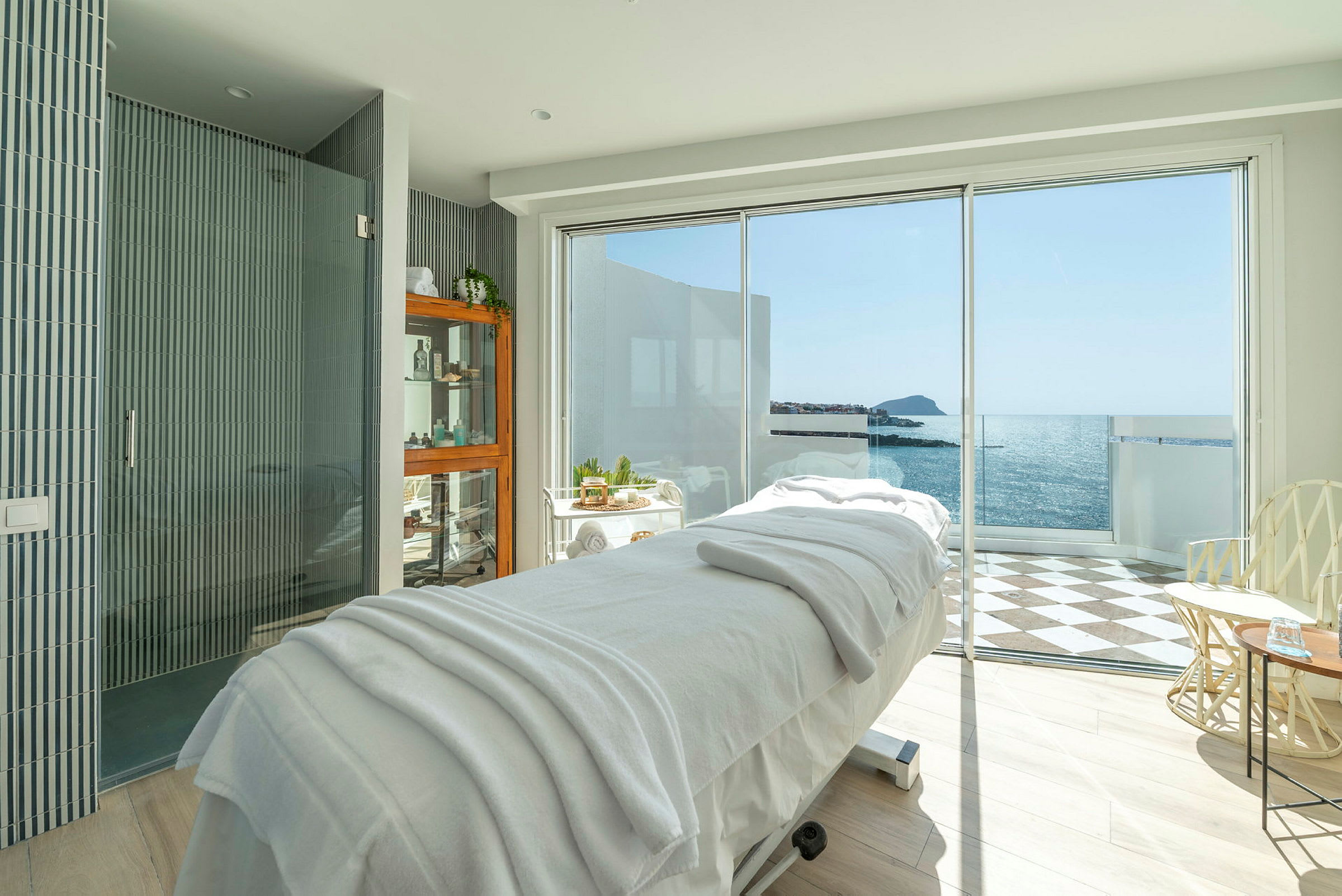 Et spa-behandlingsrum med en massagebriks foran store vinduer med udsigt til havet. (AI-genereret tekst)