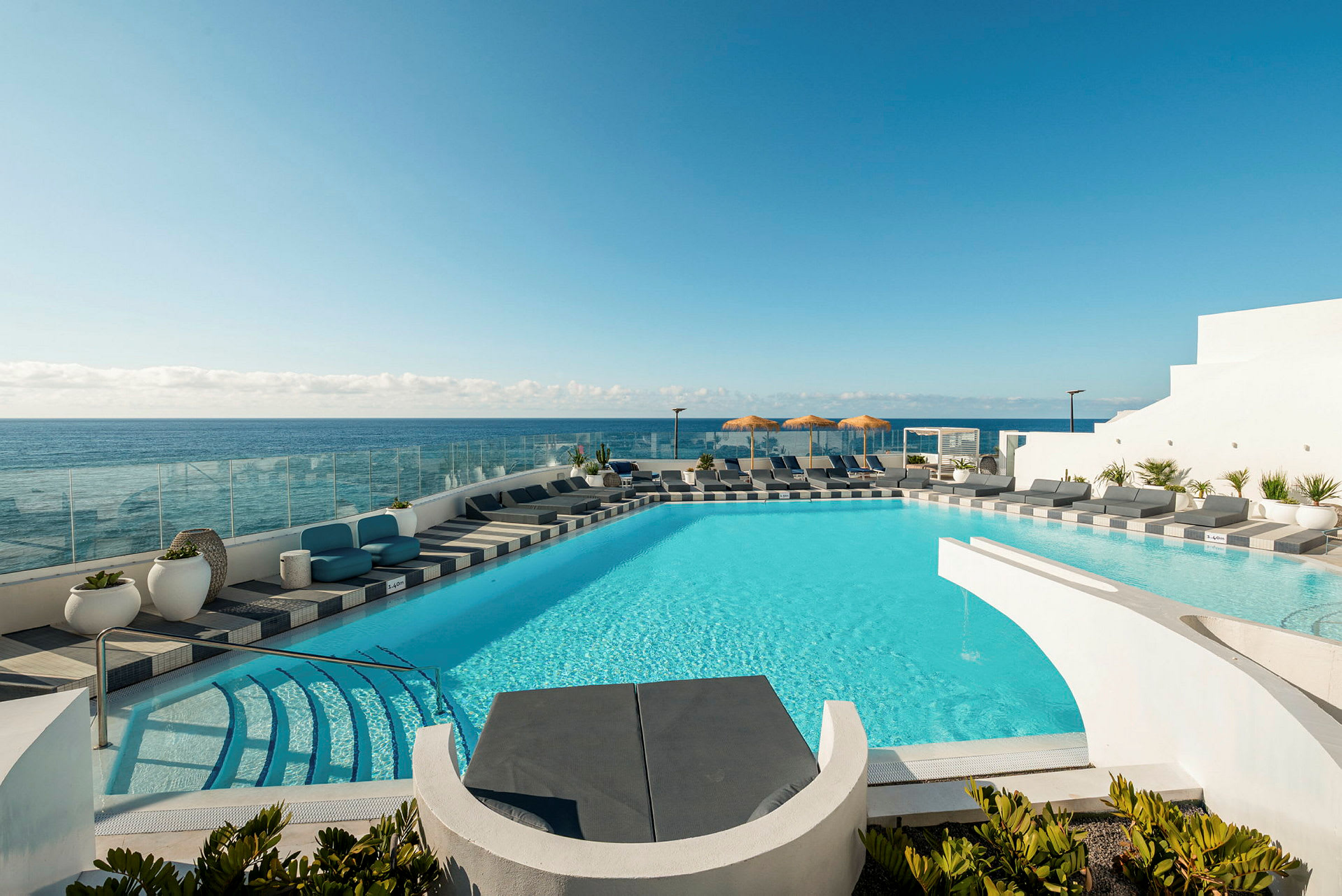 En pool med turkis vand omgivet af solsenge og udsigt over havet. I baggrunden ses en klar blå himmel. (AI-genereret tekst)