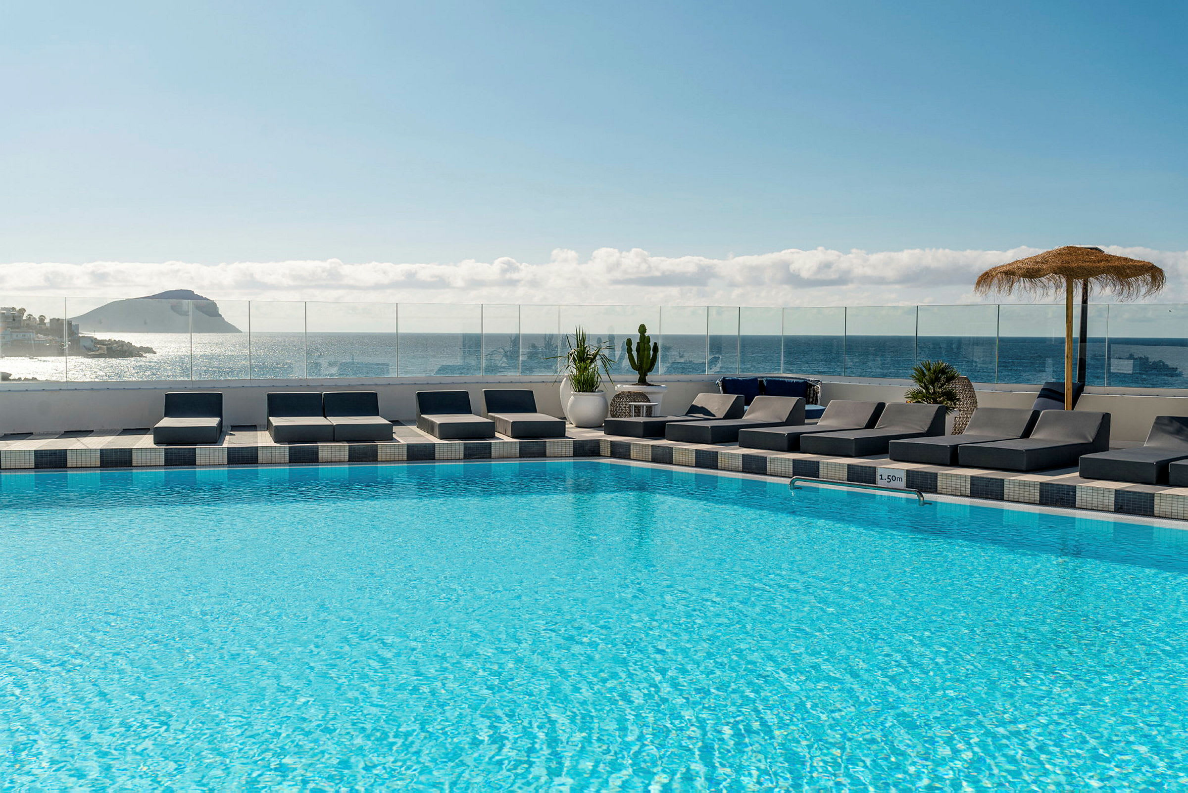 En pool på et hotel med solsenge omkring samt udsigt over havet og en ø i baggrunden. (AI-genereret tekst)