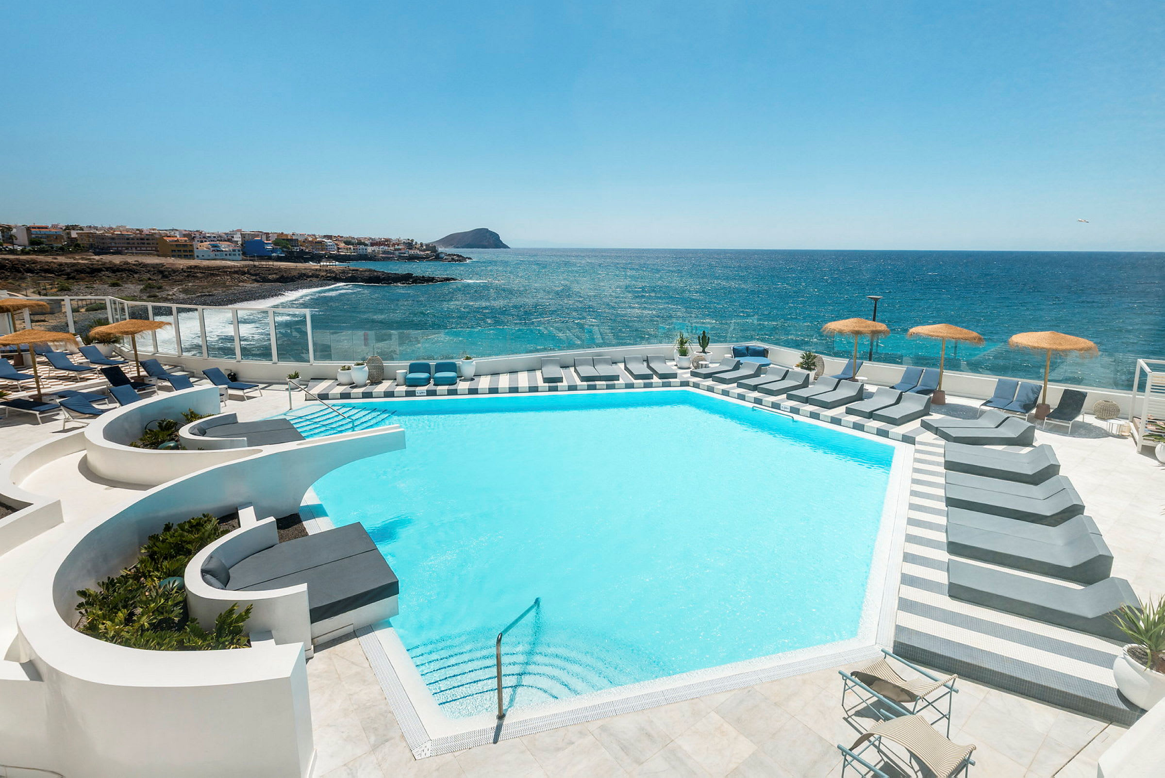 En moderne pool med liggestole og parasoller med udsigt over havet og en kystlinje i baggrunden. (AI-genereret tekst)