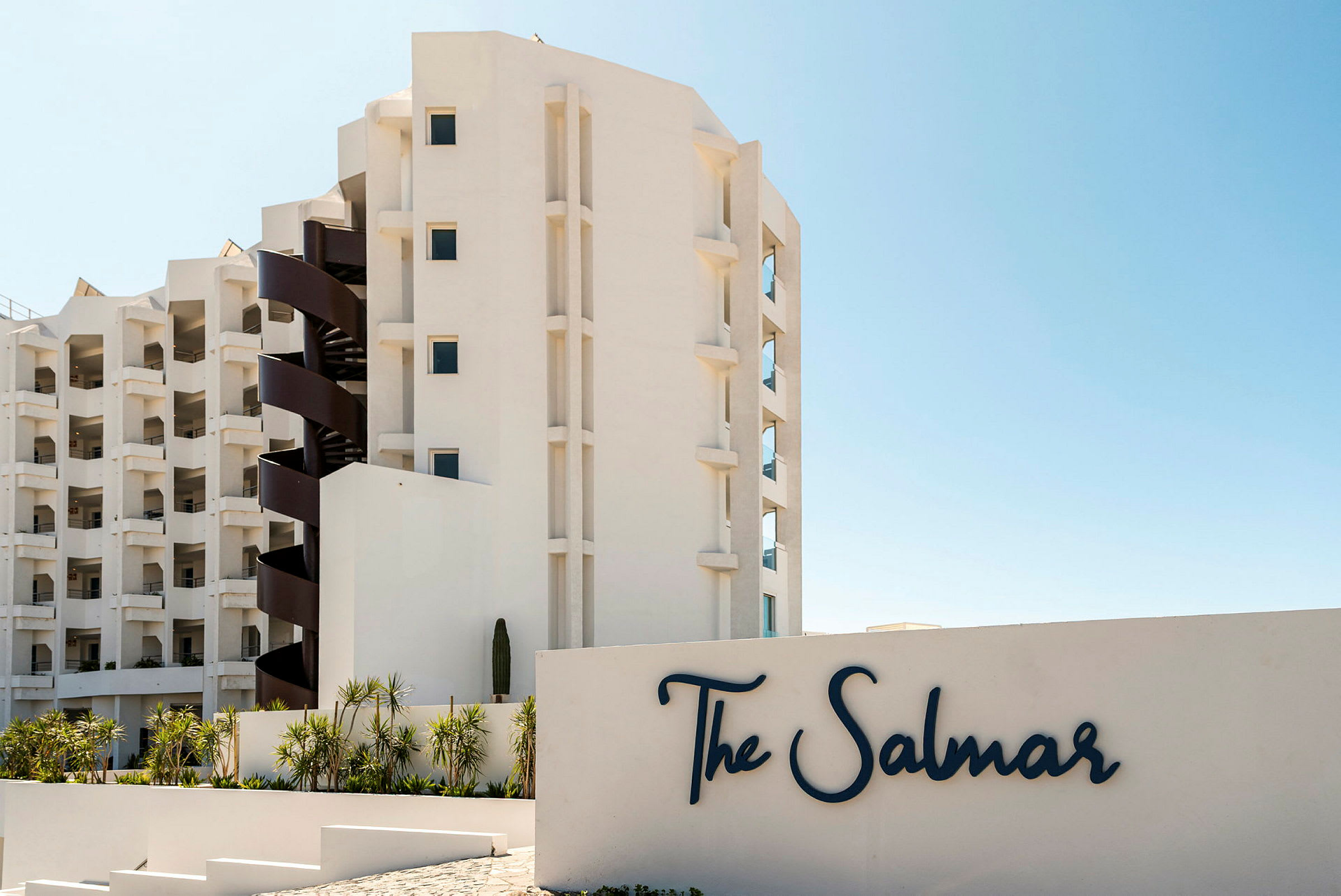 En moderne bygning med altaner og en spiraltrappe ved siden af et skilt med teksten "The Salmar". Foran bygningen er der beplantning med palmer. (AI-genereret tekst)