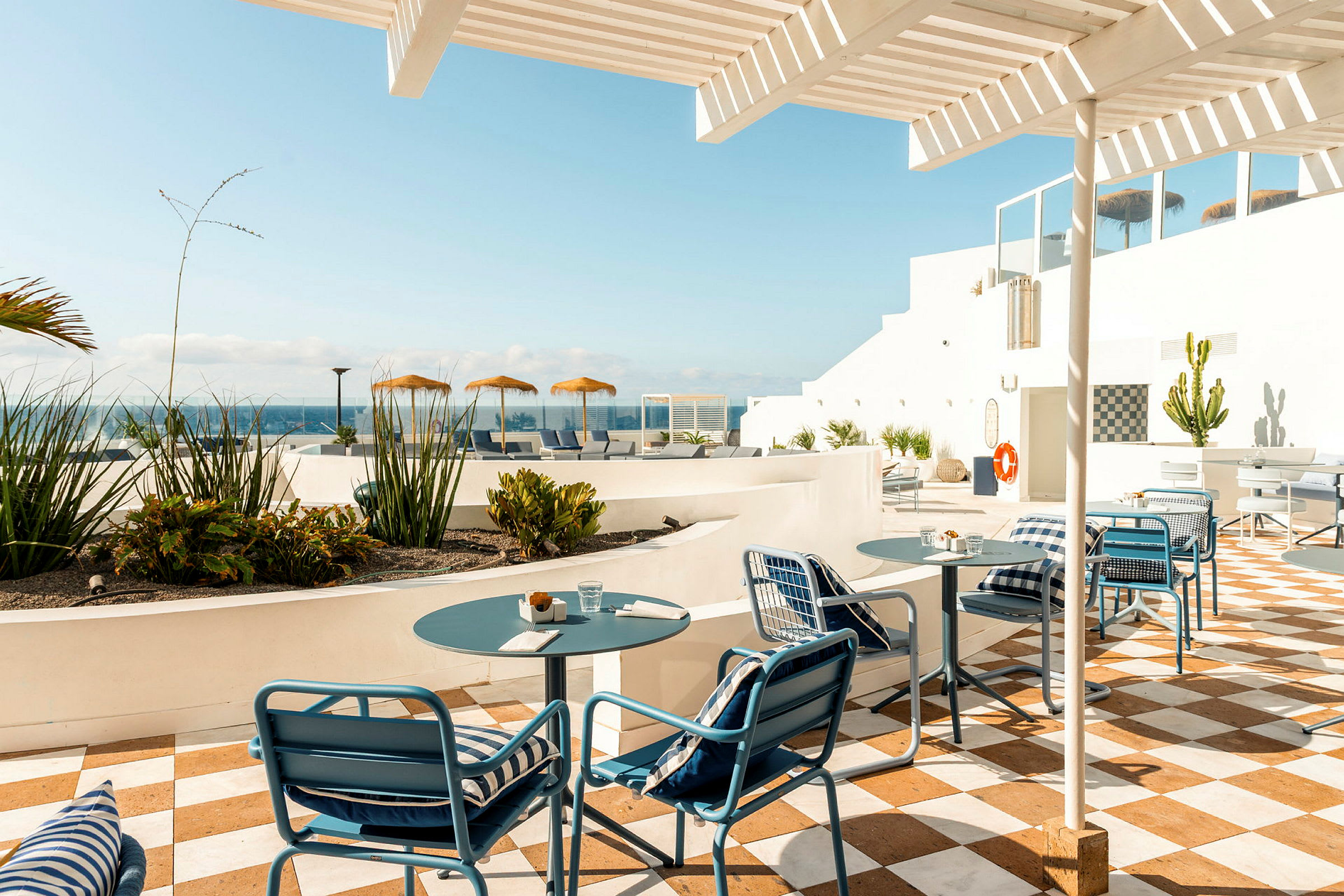 Udendørs spiseområde med borde og stole under en pergola, omgivet af grønne planter og med udsigt over havet. (AI-genereret tekst)