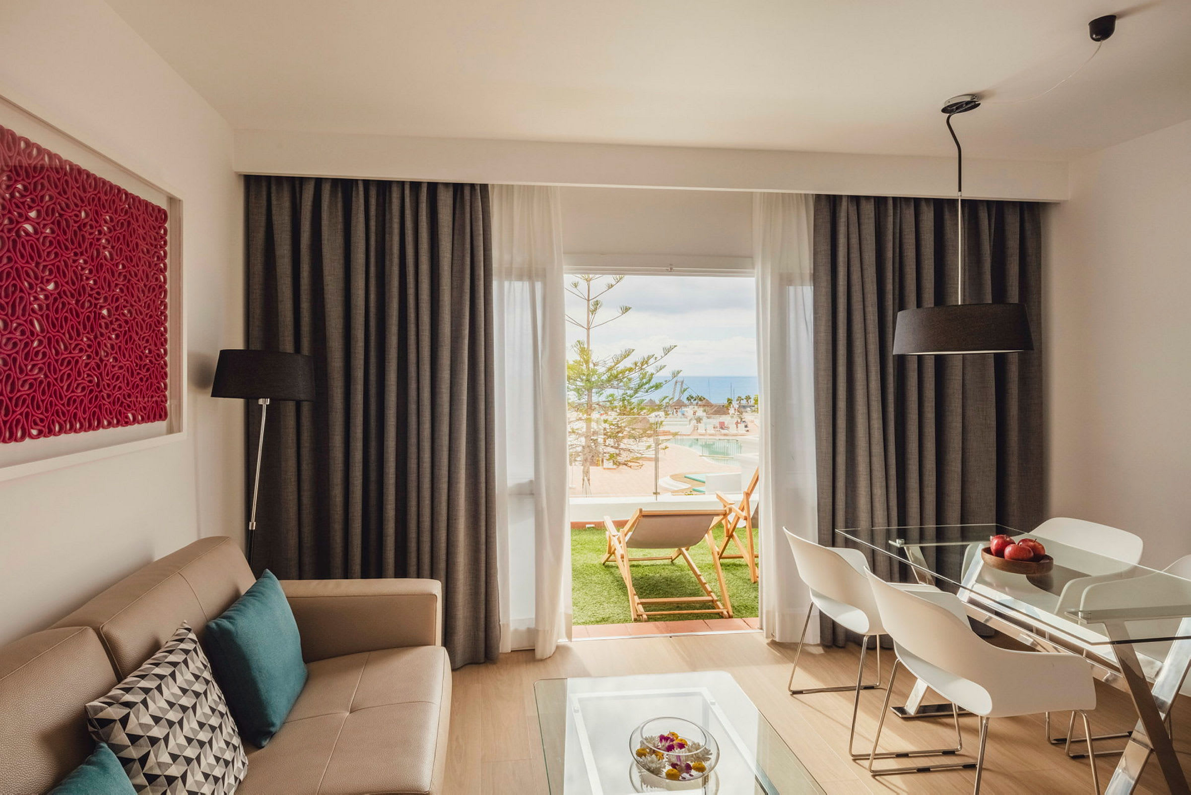 En moderne stue med en beige sofa og spisebord med hvide stole. Rummet har store vinduer med gardiner og udsigt til en grøn terrasse med liggestole og en pool. (AI-genereret tekst)