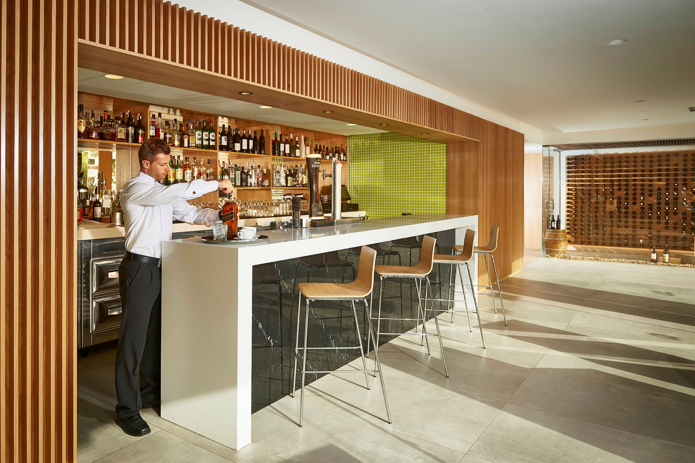 En bartender forbereder en drink ved en moderne bar med træpaneler og en grøn flisebelagt baggrund. Barstole venter ved disken. (AI-genereret tekst)
