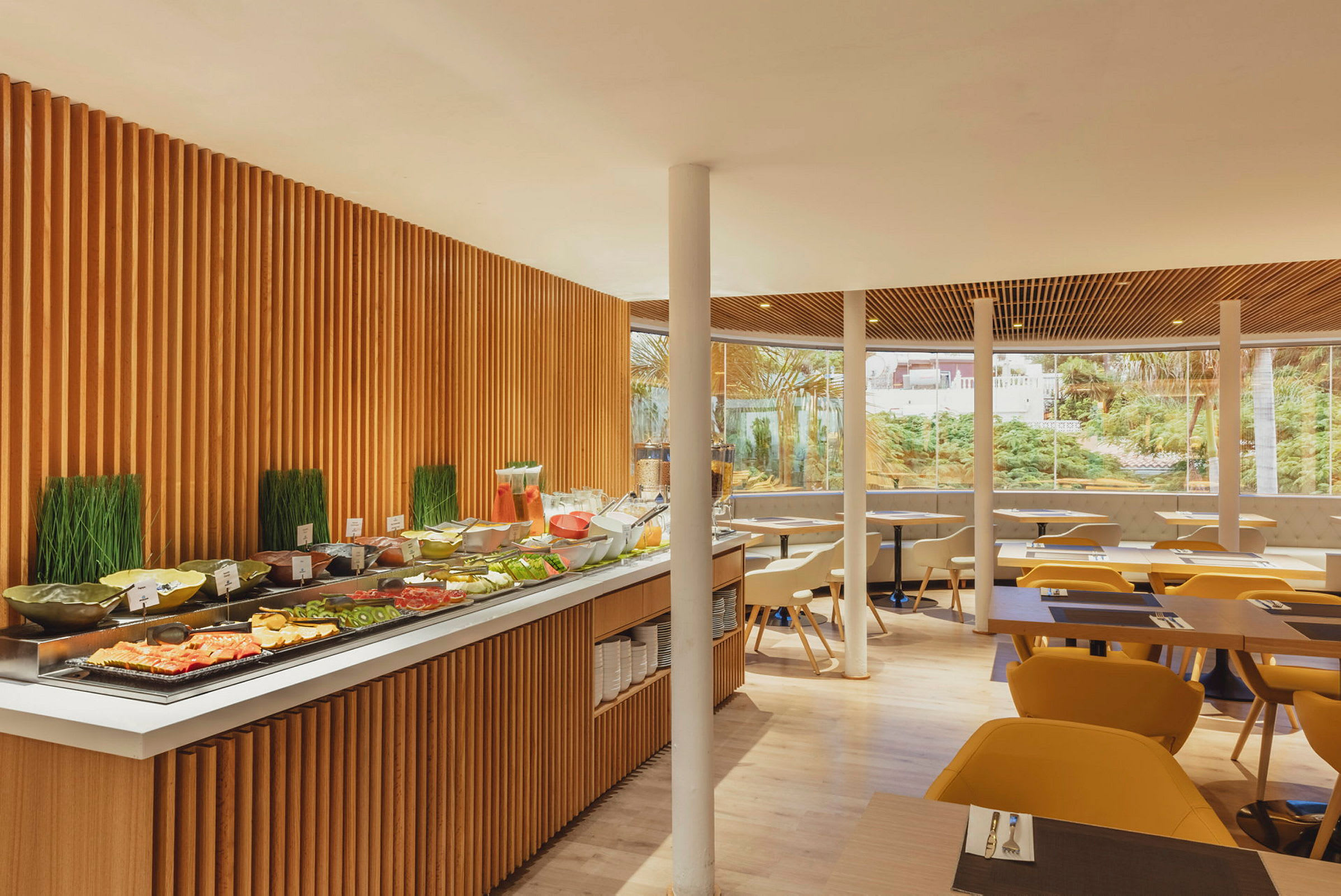 Billede af en restaurant med en buffetbord med forskellige retter og drikkevarer, omgivet af siddepladser med gulaktige stole og store vinduer, der viser en grøn have udenfor. (AI-genereret tekst)