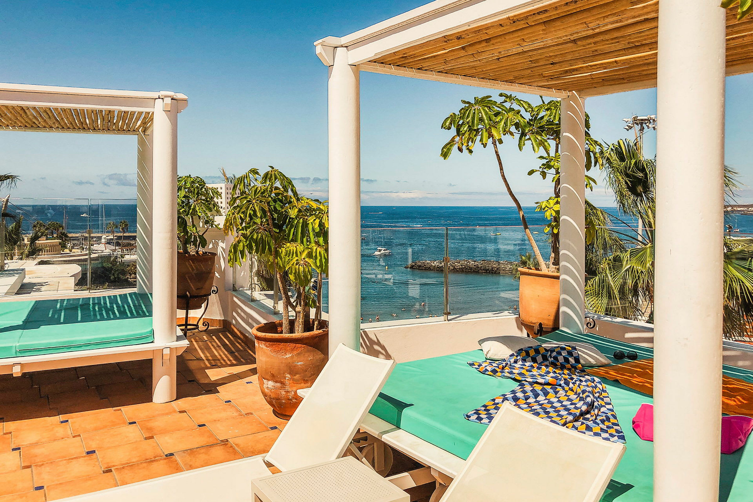 En solrig terrasse med udsigt over havet. En dagseng med turkis madras og farverige stoffer står under en pergola. Rundt terrassen er der potteplanter og palmer. (AI-genereret tekst)