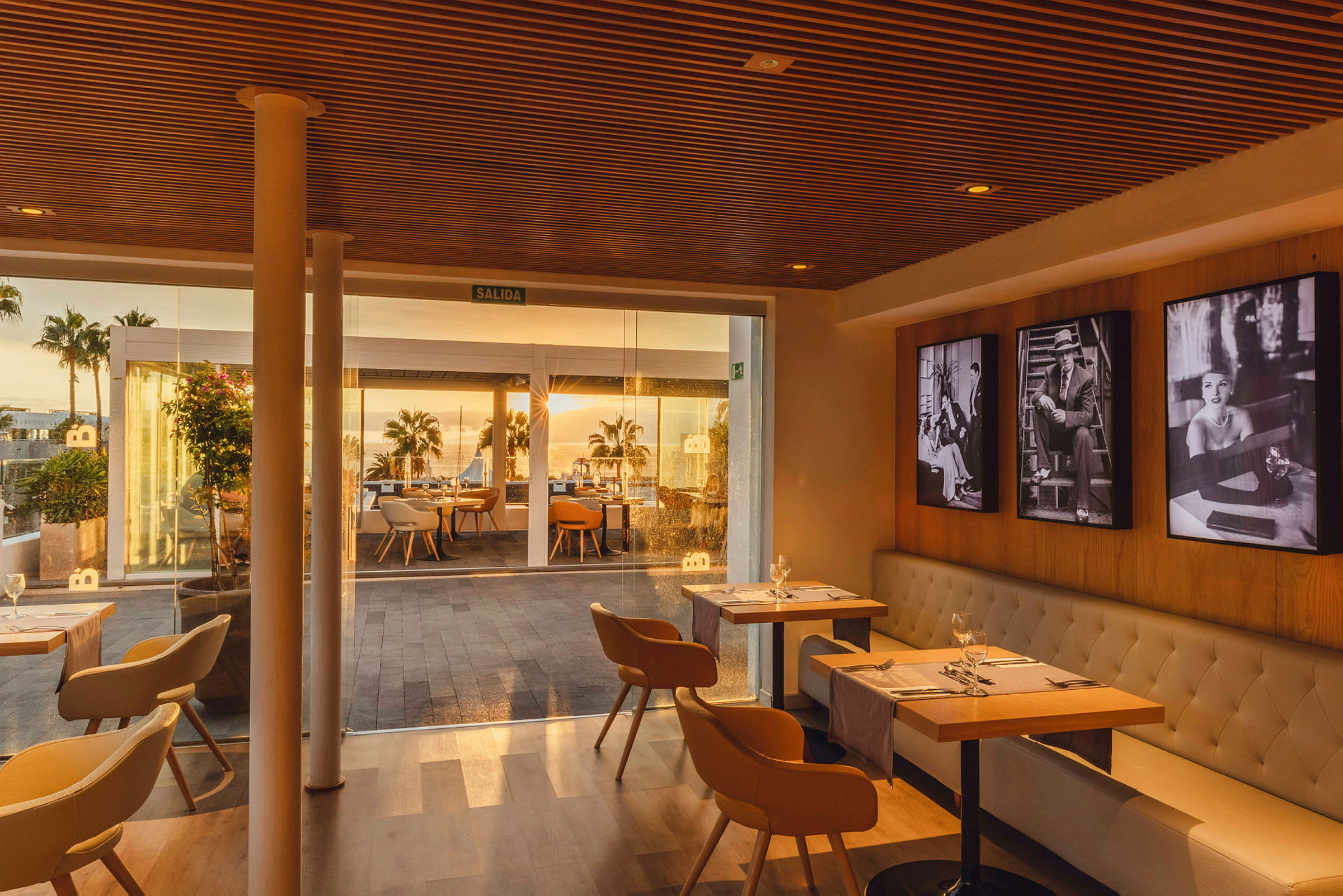 En indendørs del af en restaurant med borde og stole samt glaspartier, der viser udsigt til en udendørsterrasse med borde og palmer i solnedgang. (AI-genereret tekst)