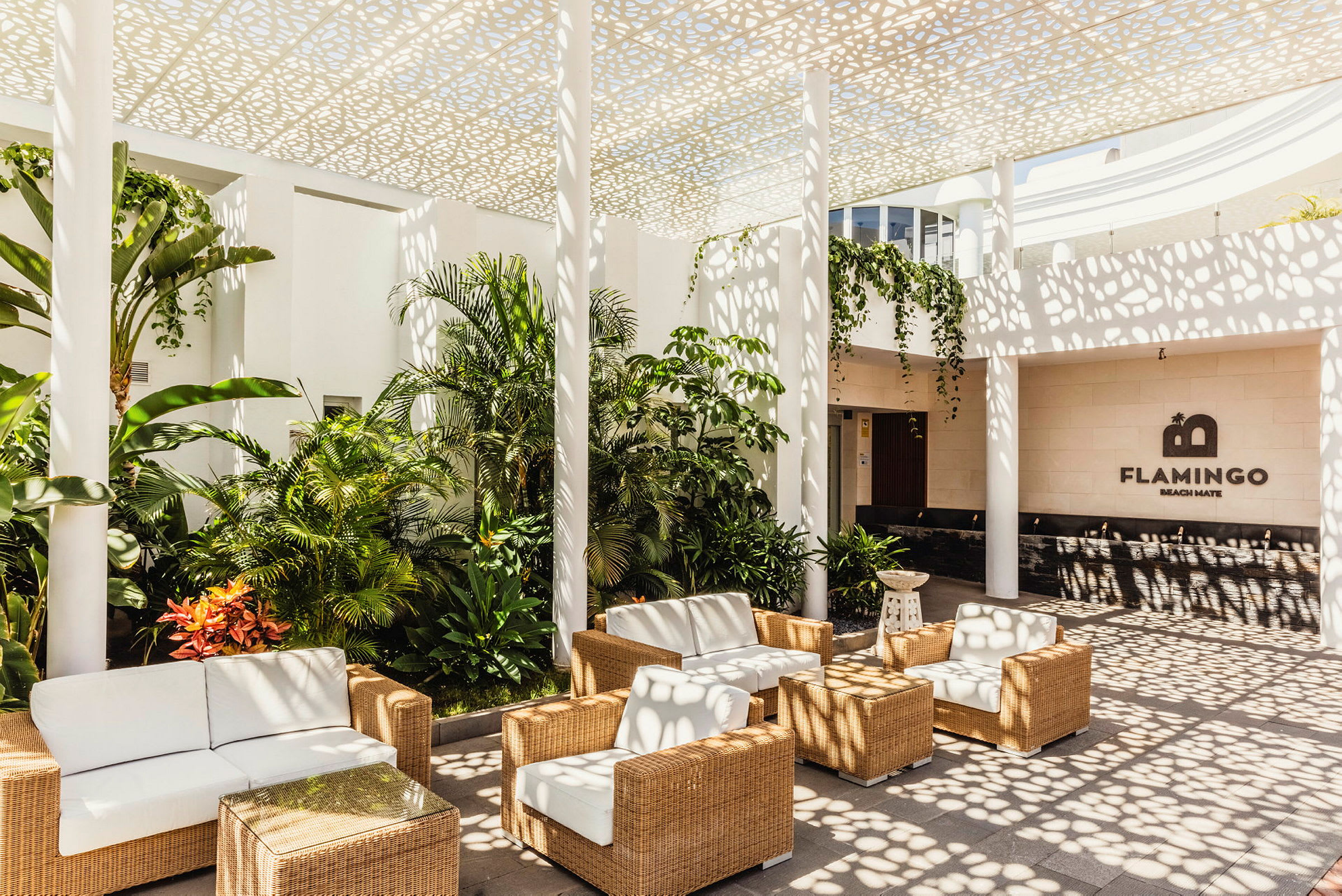 En udendørs lounge med hvide siddepladser og flettet design, omgivet af frodige planter under et mønstret loft. (AI-genereret tekst)