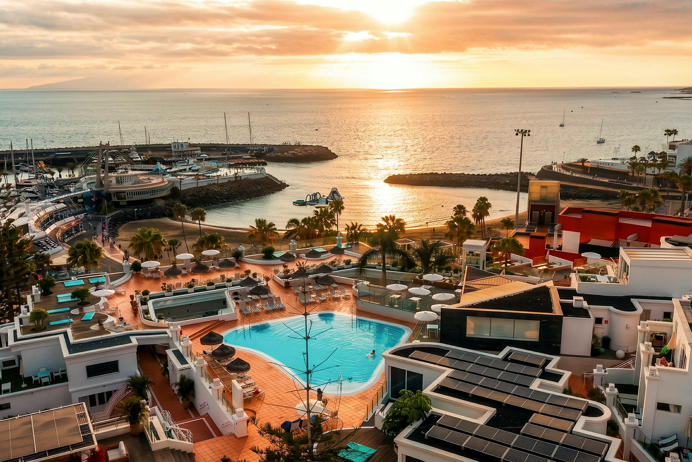 Udsigt over et resort med swimmingpools, liggestole og parasoller, med en strand og lystbådehavn ved kysten. Solnedgangens lys reflekteres i vandet. (AI-genereret tekst)