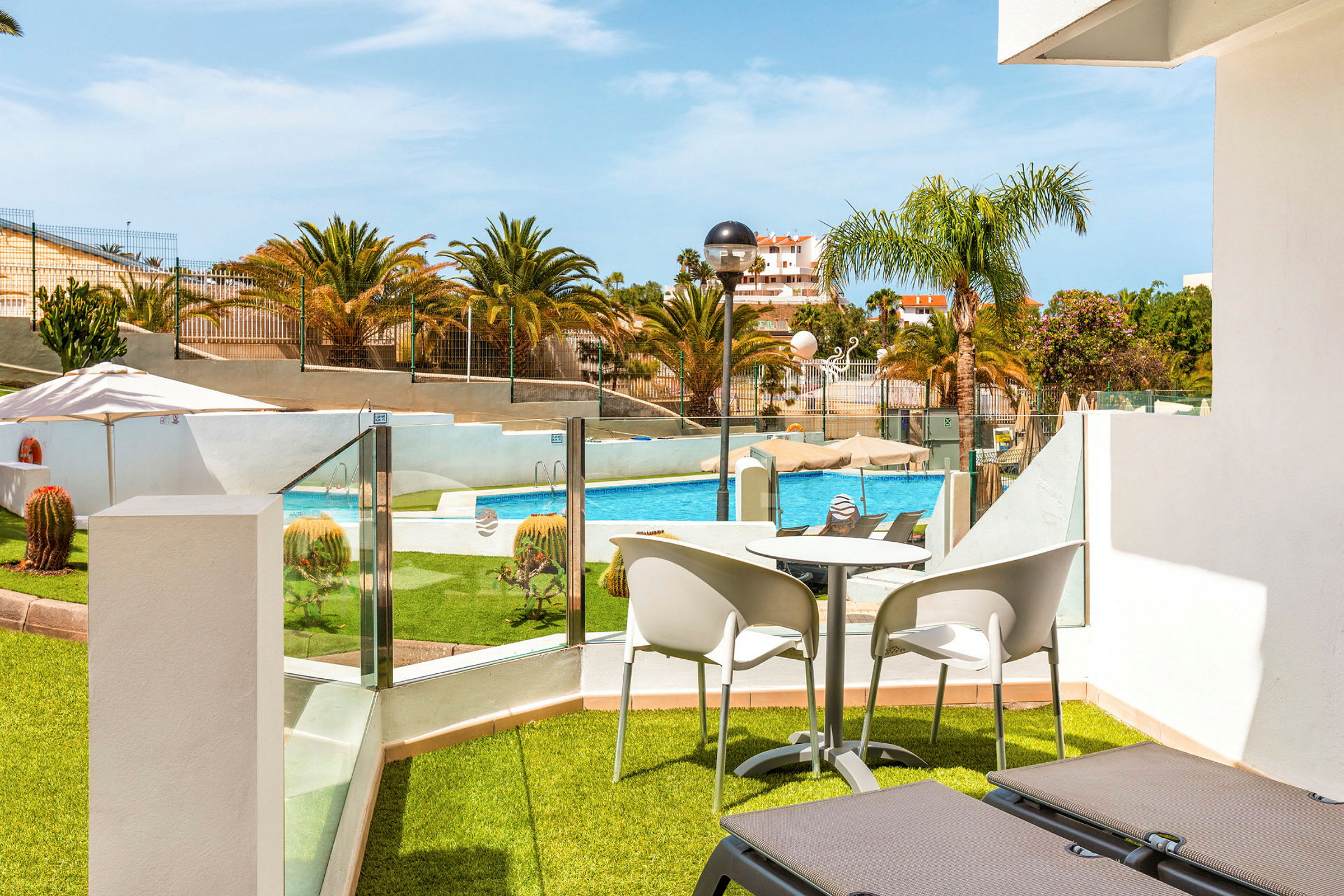 Udsigt fra en terrasse med liggestole, bord og stole mod en pool og omkringliggende grøntområder. (AI-genereret tekst)
