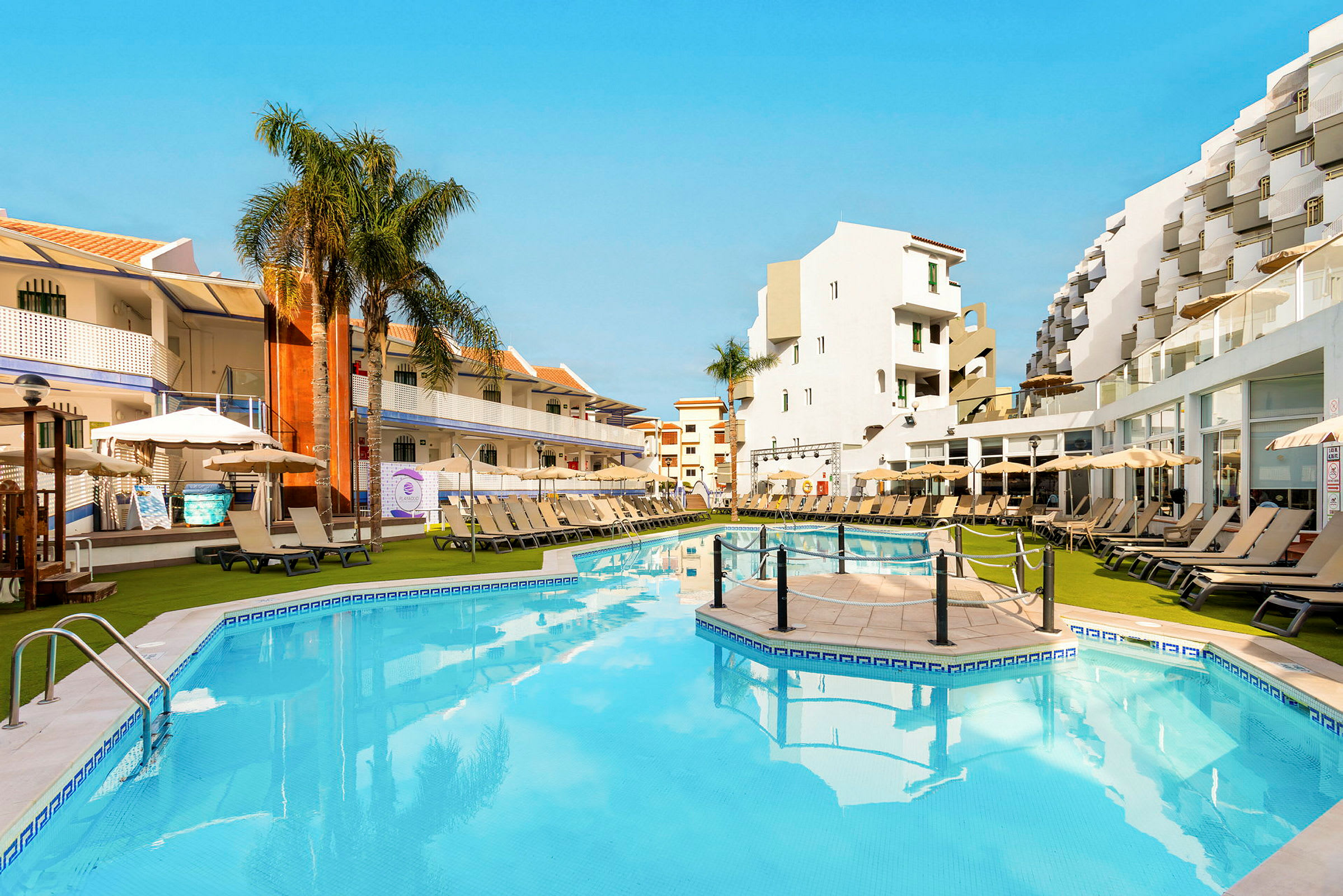 Dejlige poolområde på et hotel med liggestole, parasoller og omkringliggende bygninger. (AI-genereret tekst)