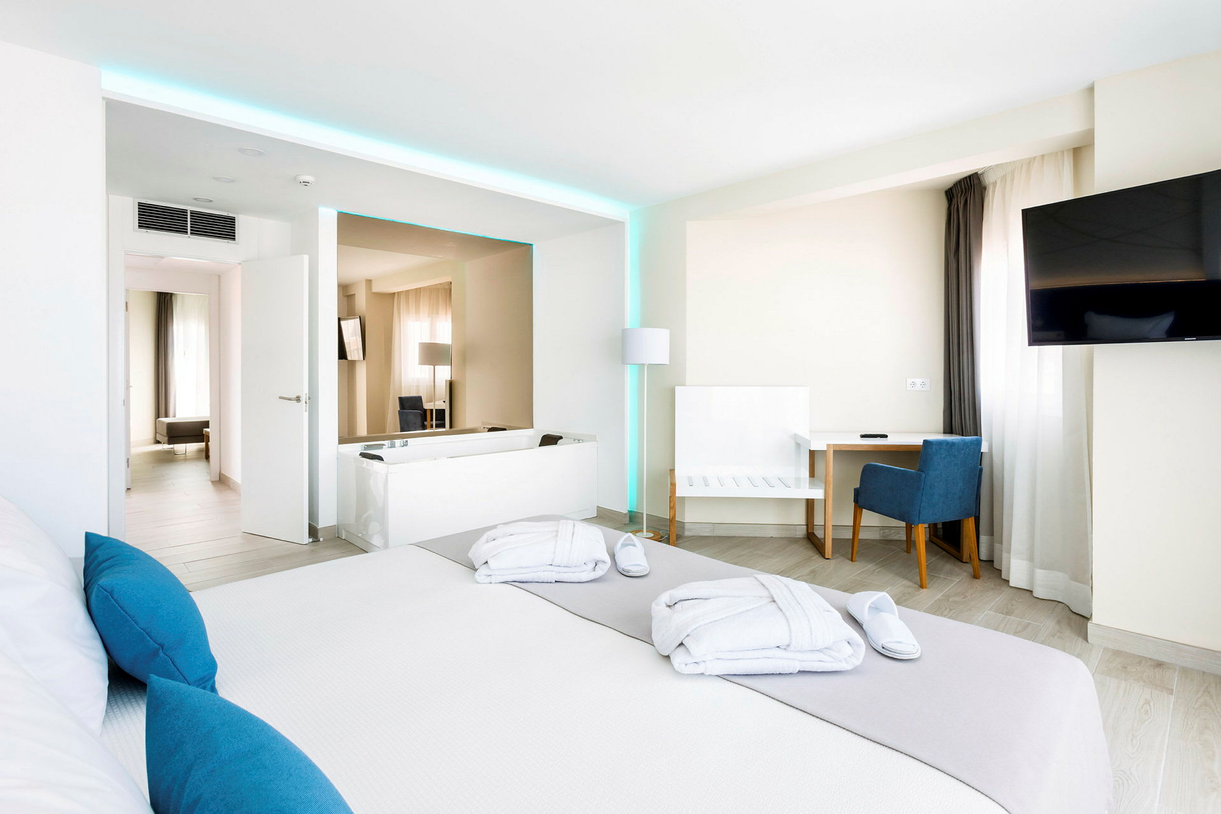 Et moderne hotelværelse med en stor seng, skrivebord og stol, et TV på væggen samt et badekar. Lyse vægge og indretning med blå accenter. (AI-genereret tekst)