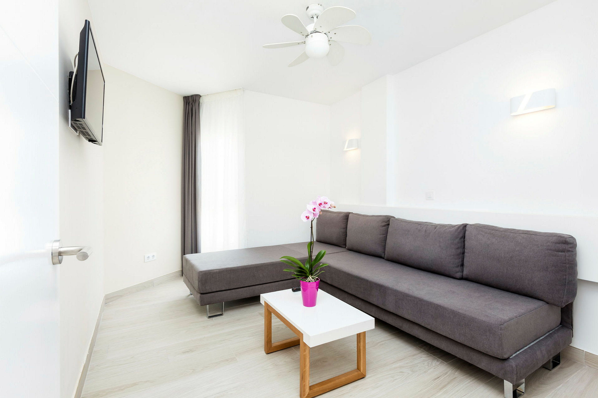 En moderne lounge med en grå sofa, hvide vægge og et lille bord med en lyserød blomsterpotte. (AI-genereret tekst)