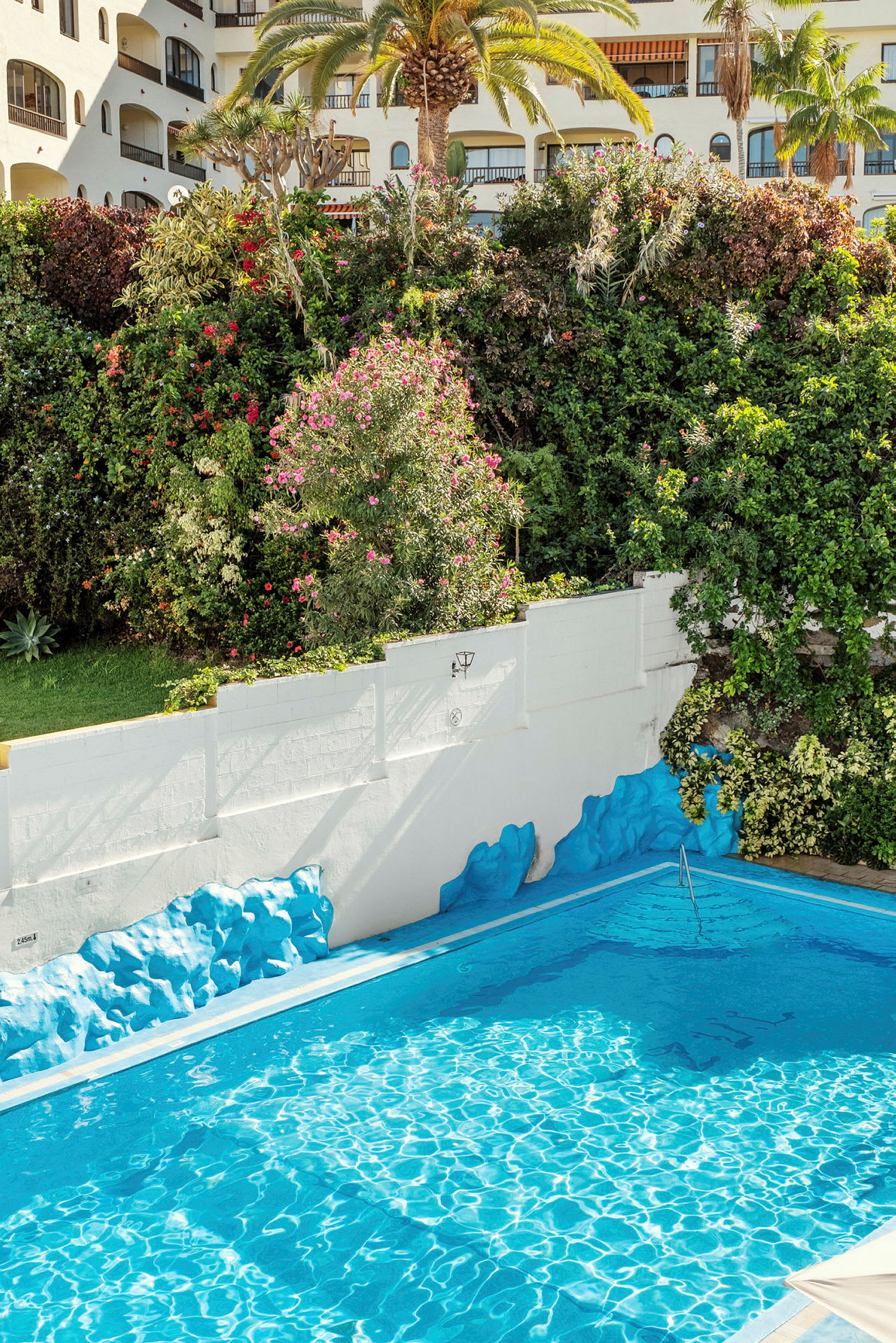 Udendørs swimmingpool med blåt vand omgivet af frodig vegetation og bygninger. (AI-genereret tekst)