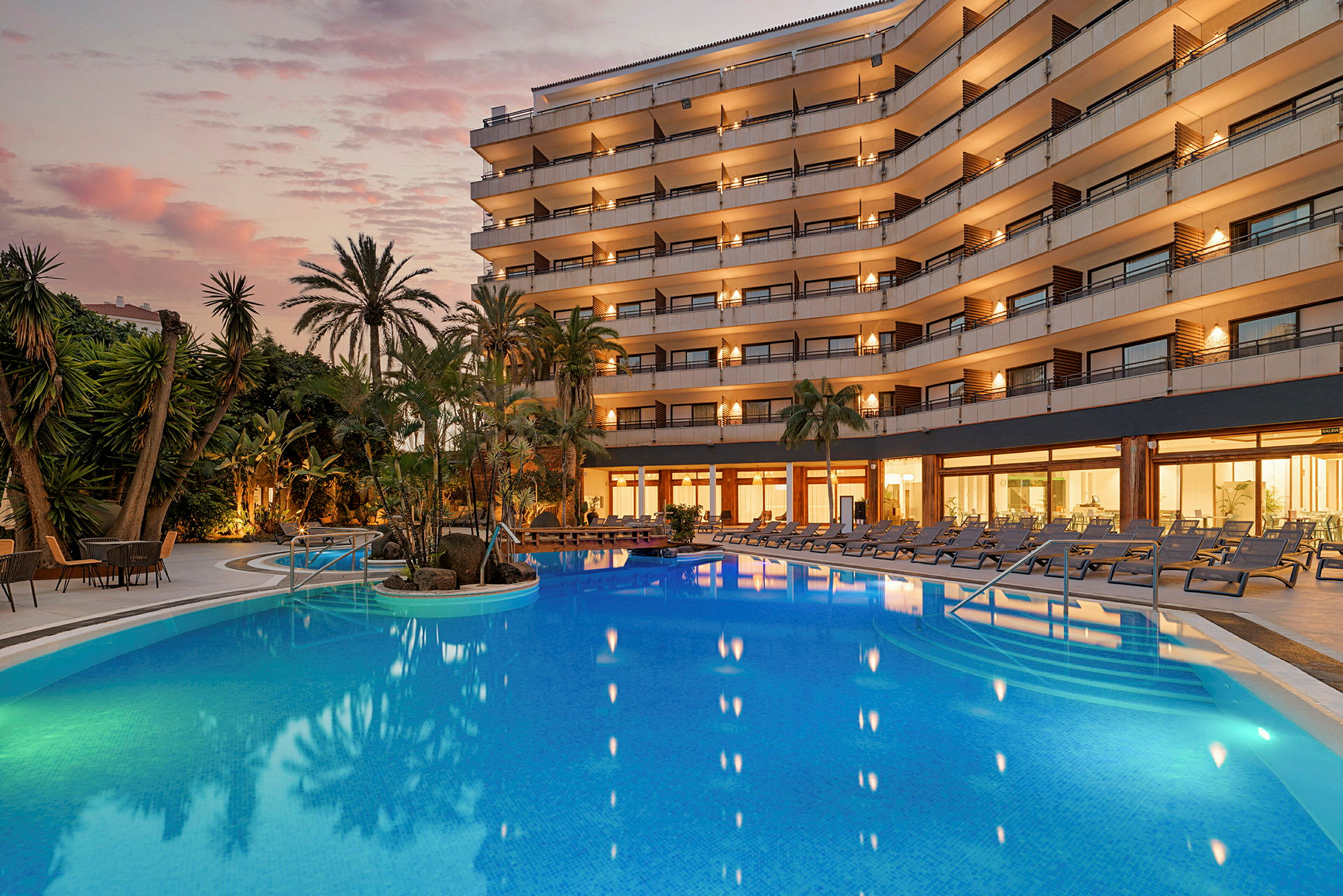 Billede af et hotel med altaner og et oplyst poolområde under skumringen. (AI-genereret tekst)