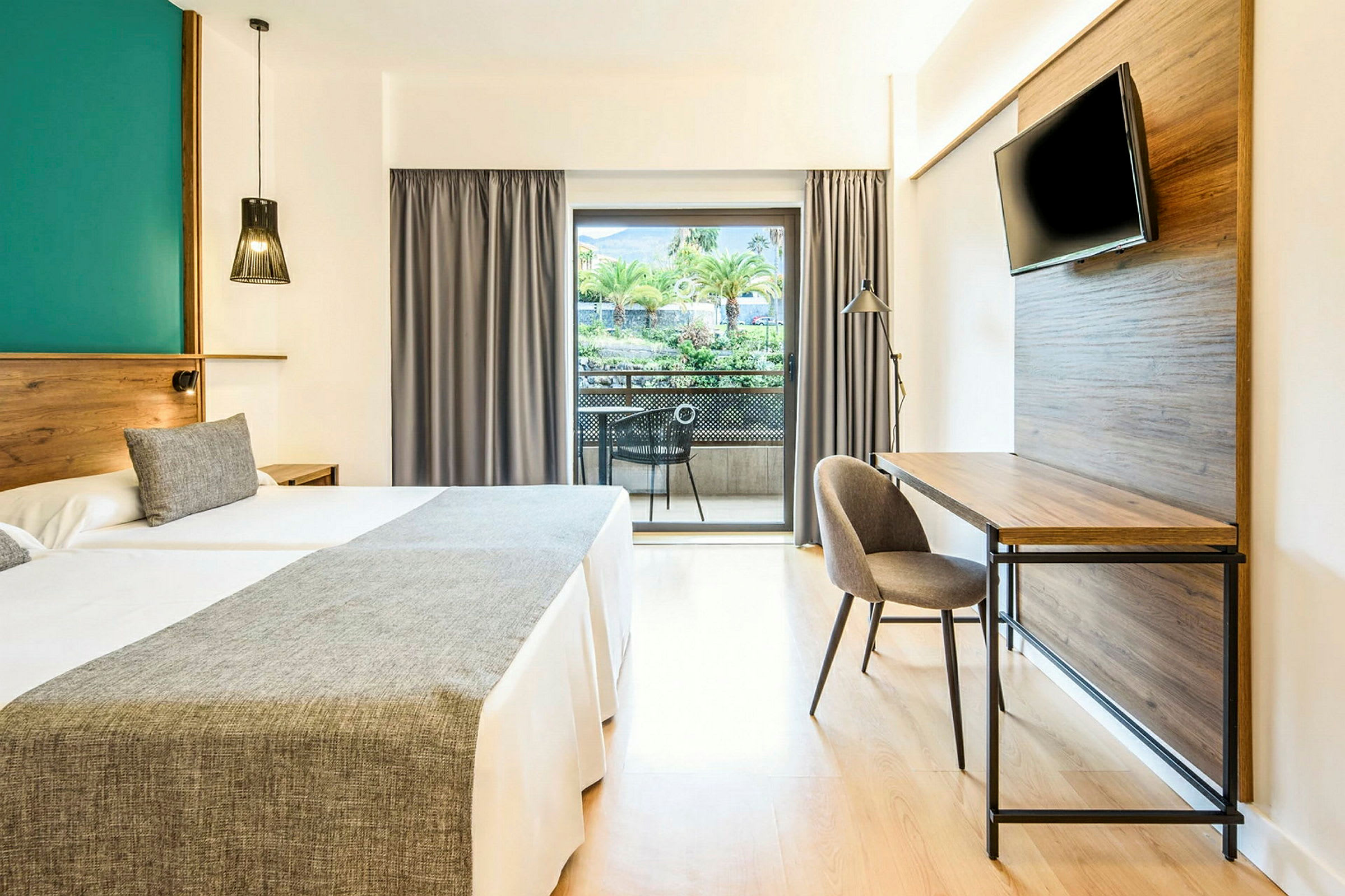Hotelværelse med to senge, skrivebord, tv og balkon med udsigt over træer og grønne områder. (AI-genereret tekst)