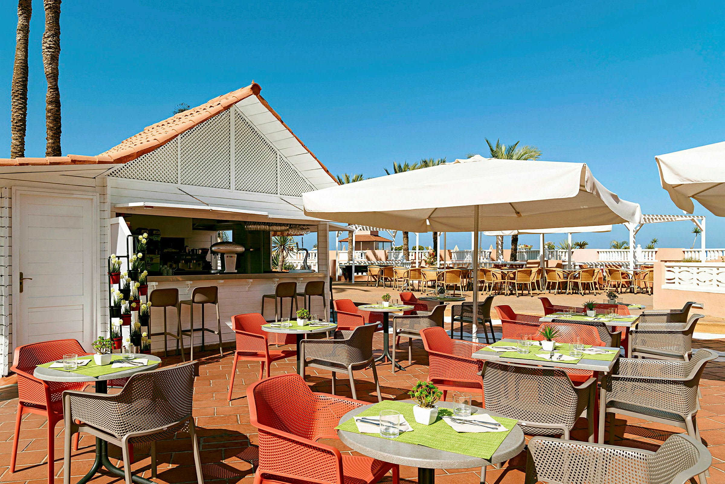 En udendørs restaurant med borde og farvestrålende stole samt parasoller. En bar i baggrunden. (AI-genereret tekst)