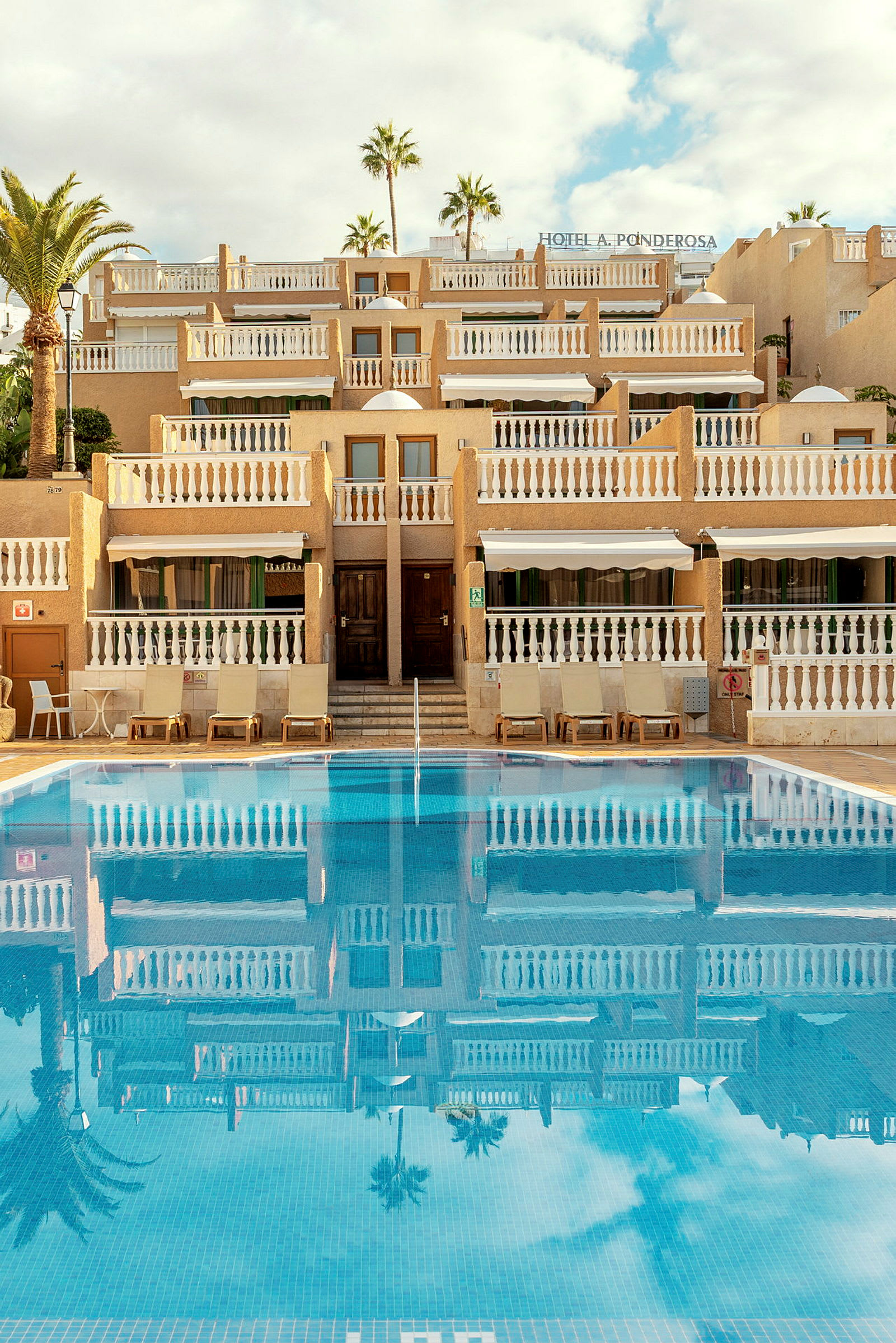Billede af et hotel med flere etager og symmetriske altaner ved siden af en klarblå pool. (AI-genereret tekst)