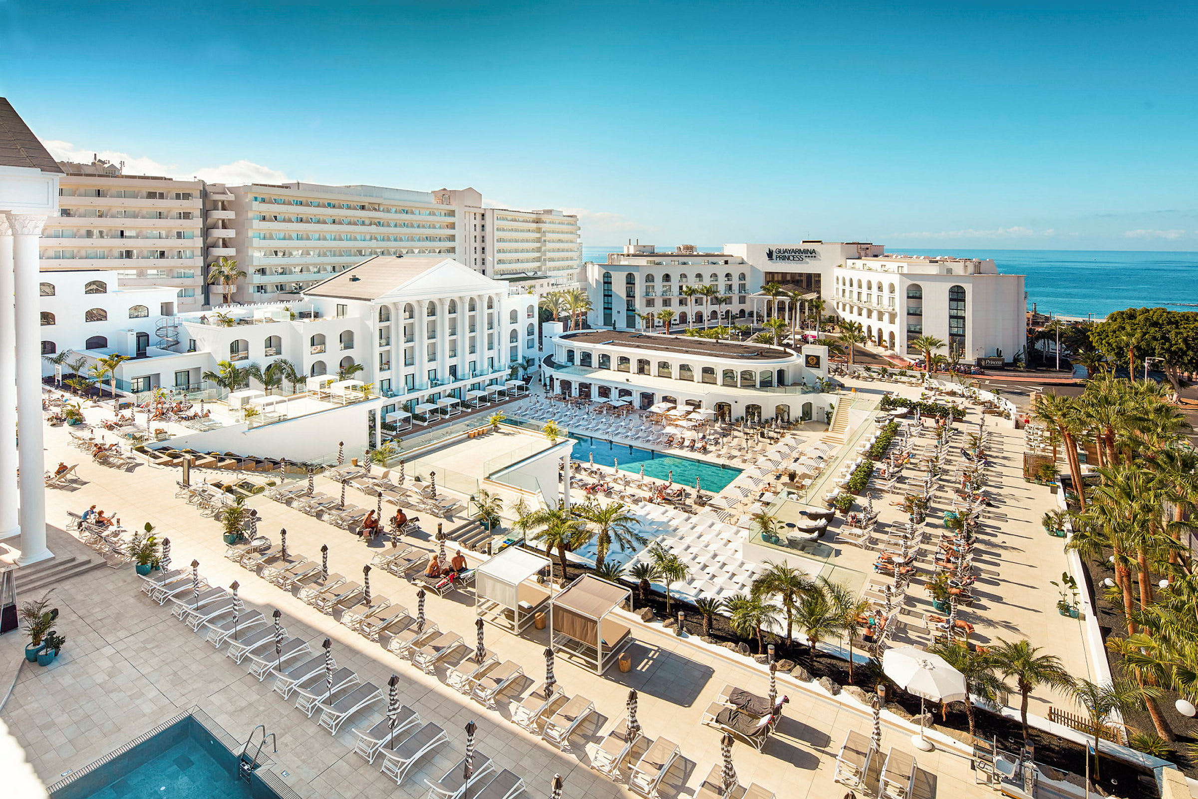 Udsigt over et hotelområde med pools, solsenge og hvid arkitektur tæt på havet. (AI-genereret tekst)