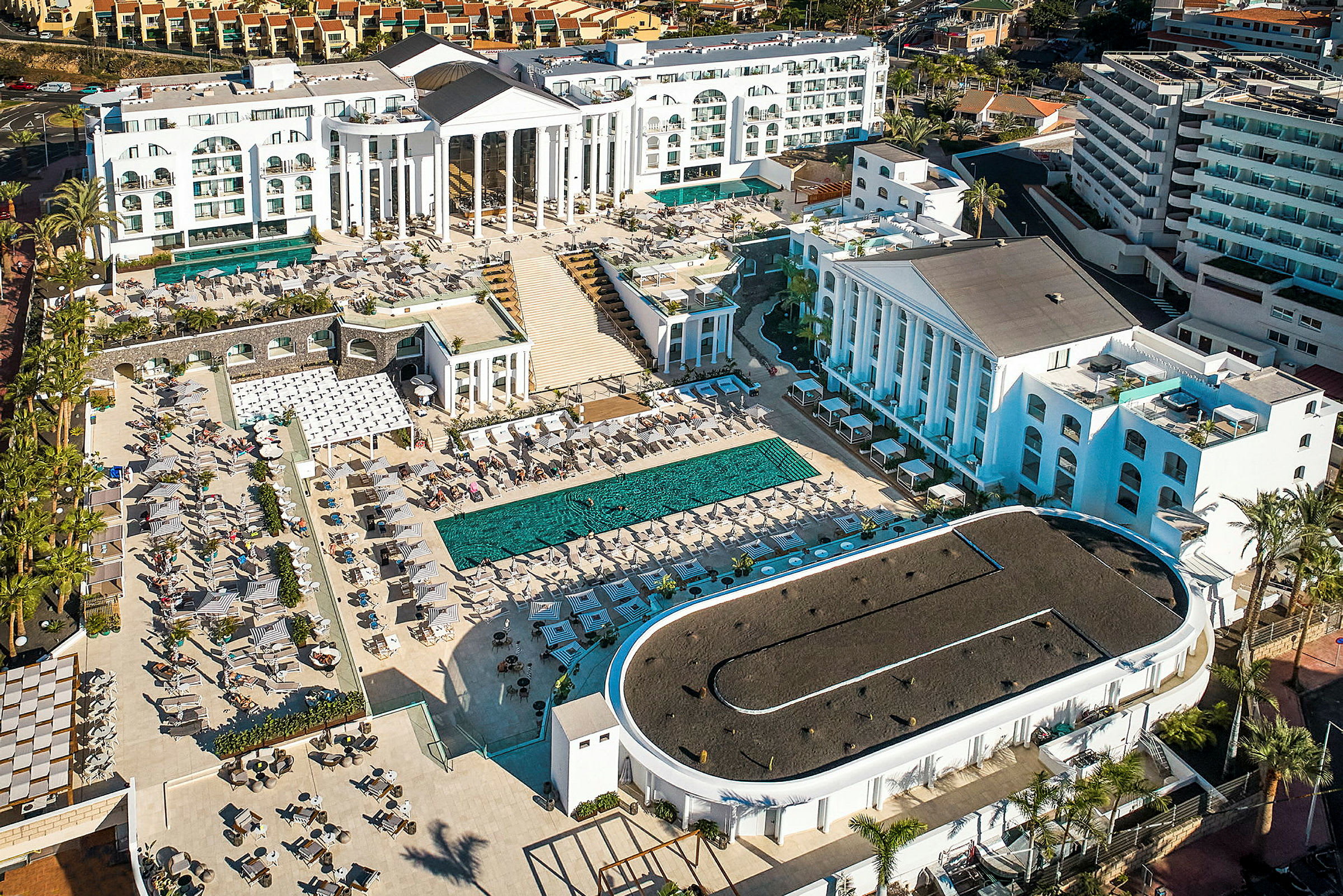 Luftfoto af et større hotelkompleks med swimmingpool og liggestole. (AI-genereret tekst)