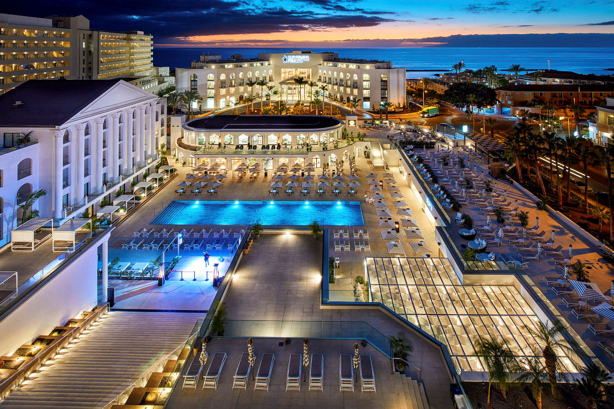 En udsigt over et hotelområde med pools og liggestole, omgivet af bygninger ved havet. (AI-genereret tekst)