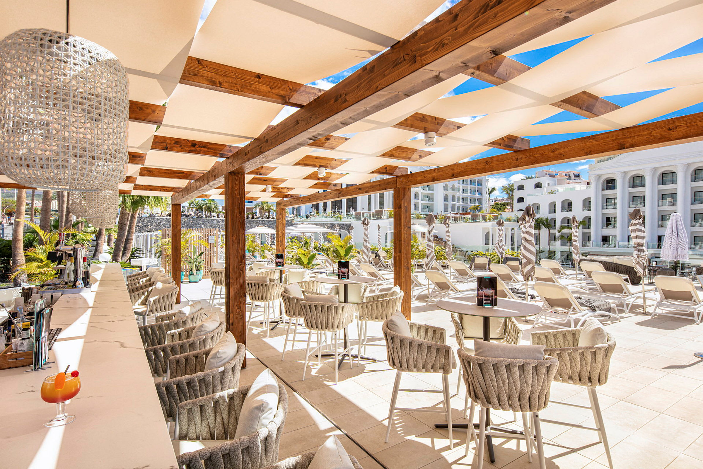 En udendørsbar med stole og borde under en pergola, omgivet af liggestole og grønt. (AI-genereret tekst)