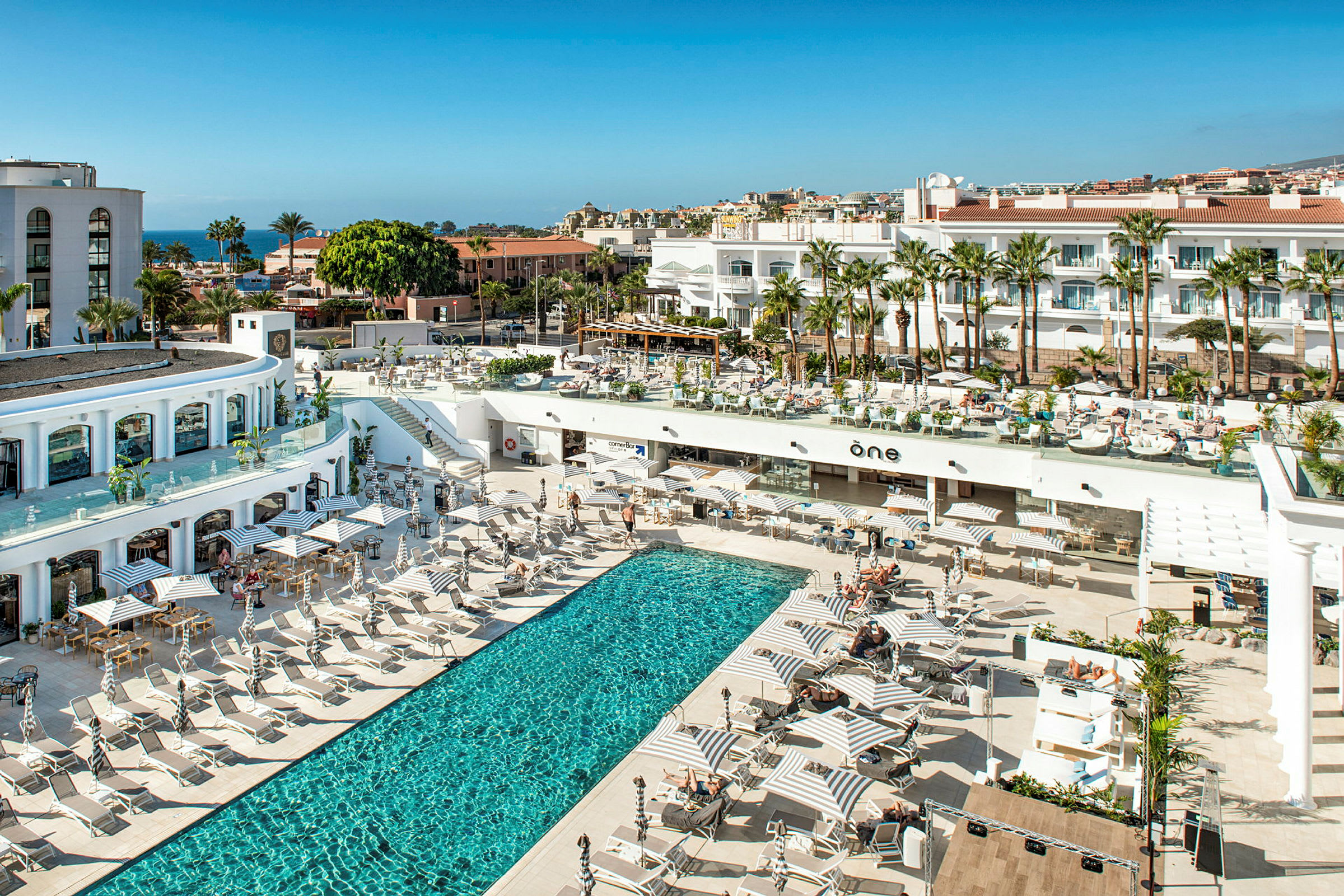 Billede af et hotelområde med en udendørs pool, liggestole og parasoller, omkranset af bygninger og palmer. (AI-genereret tekst)