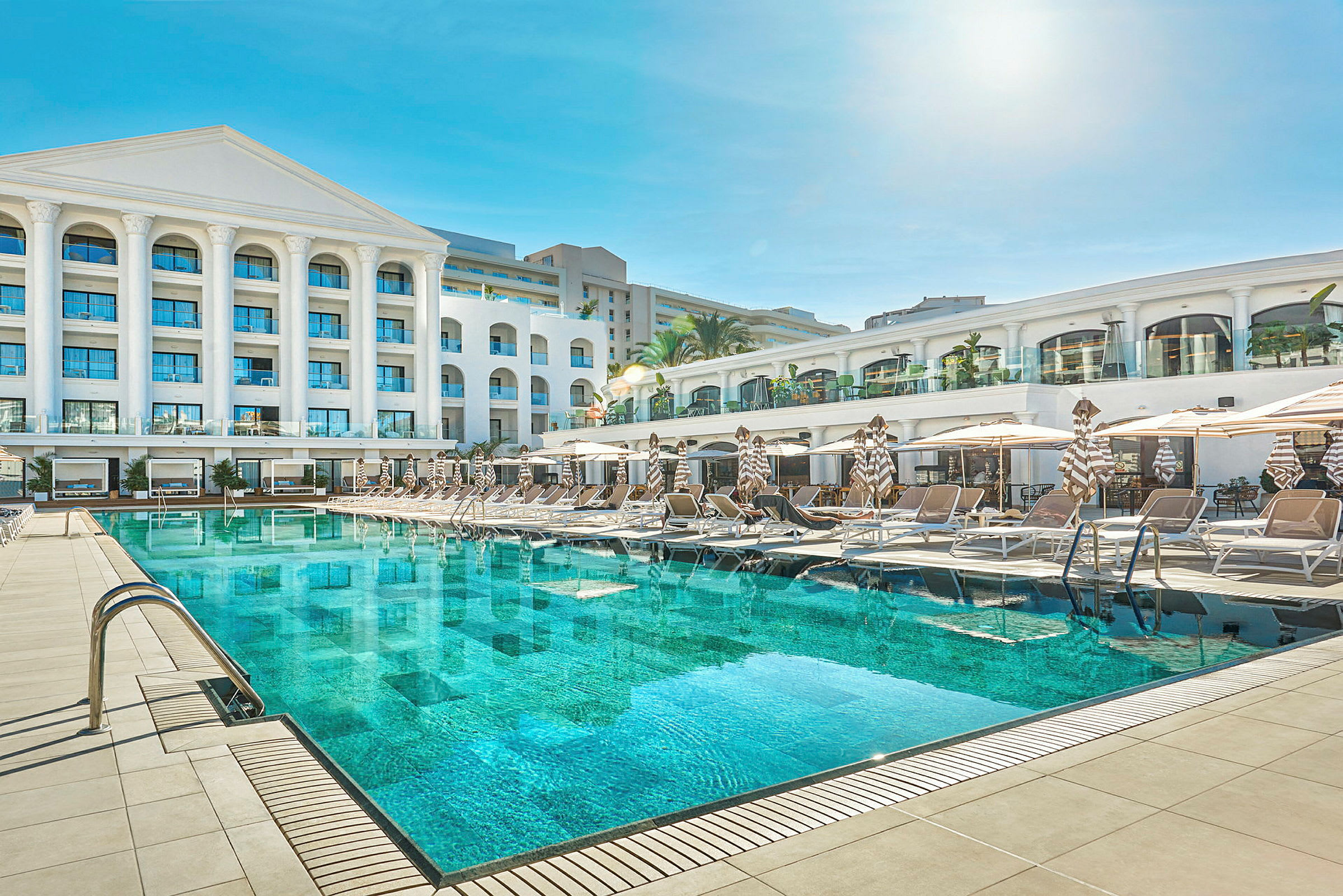 En hotelbygning med en stor swimmingpool og liggestole foran. (AI-genereret tekst)