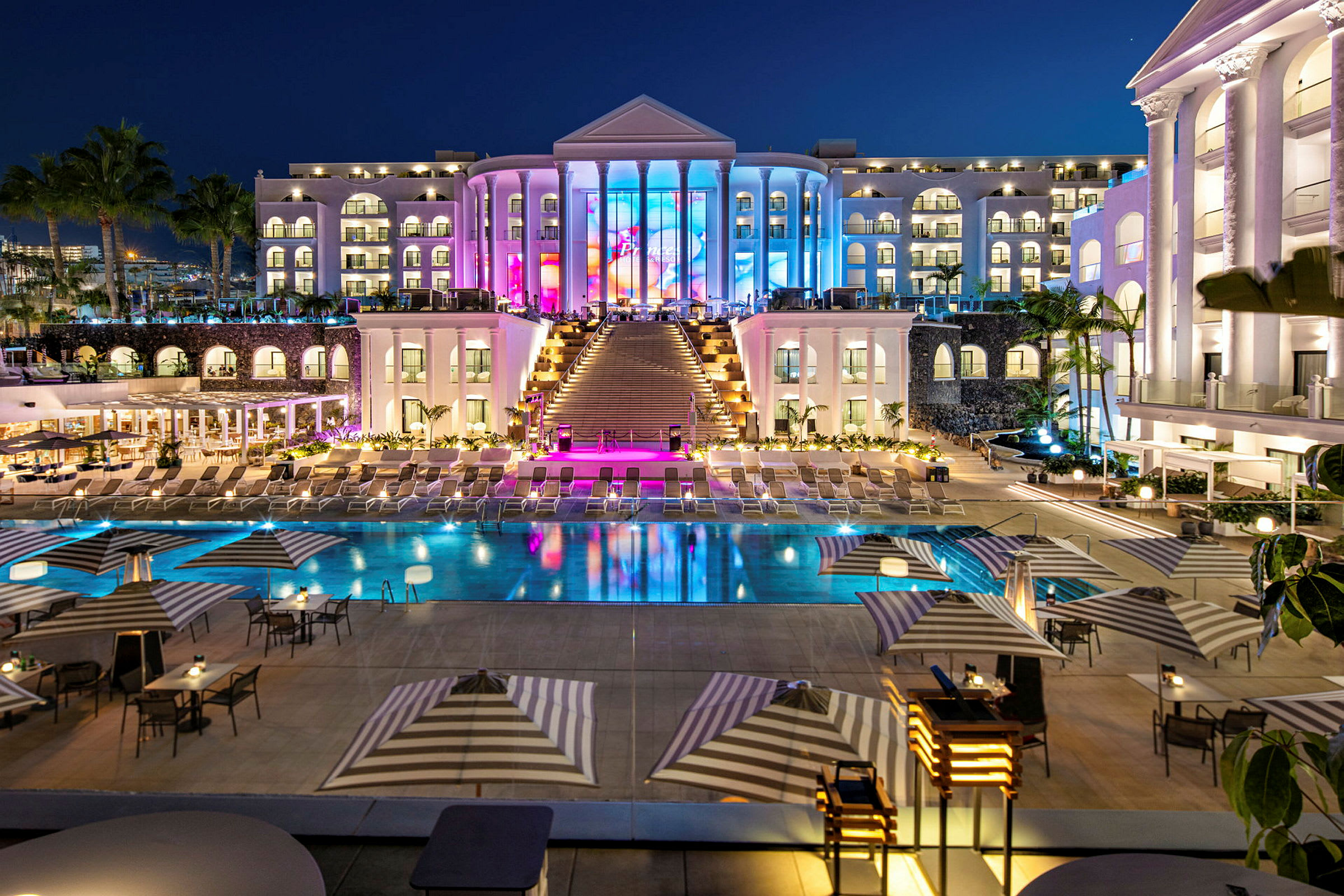 Eksklusivt hotel med oplyst facade, pool og liggestole i forgrunden. (AI-genereret tekst)