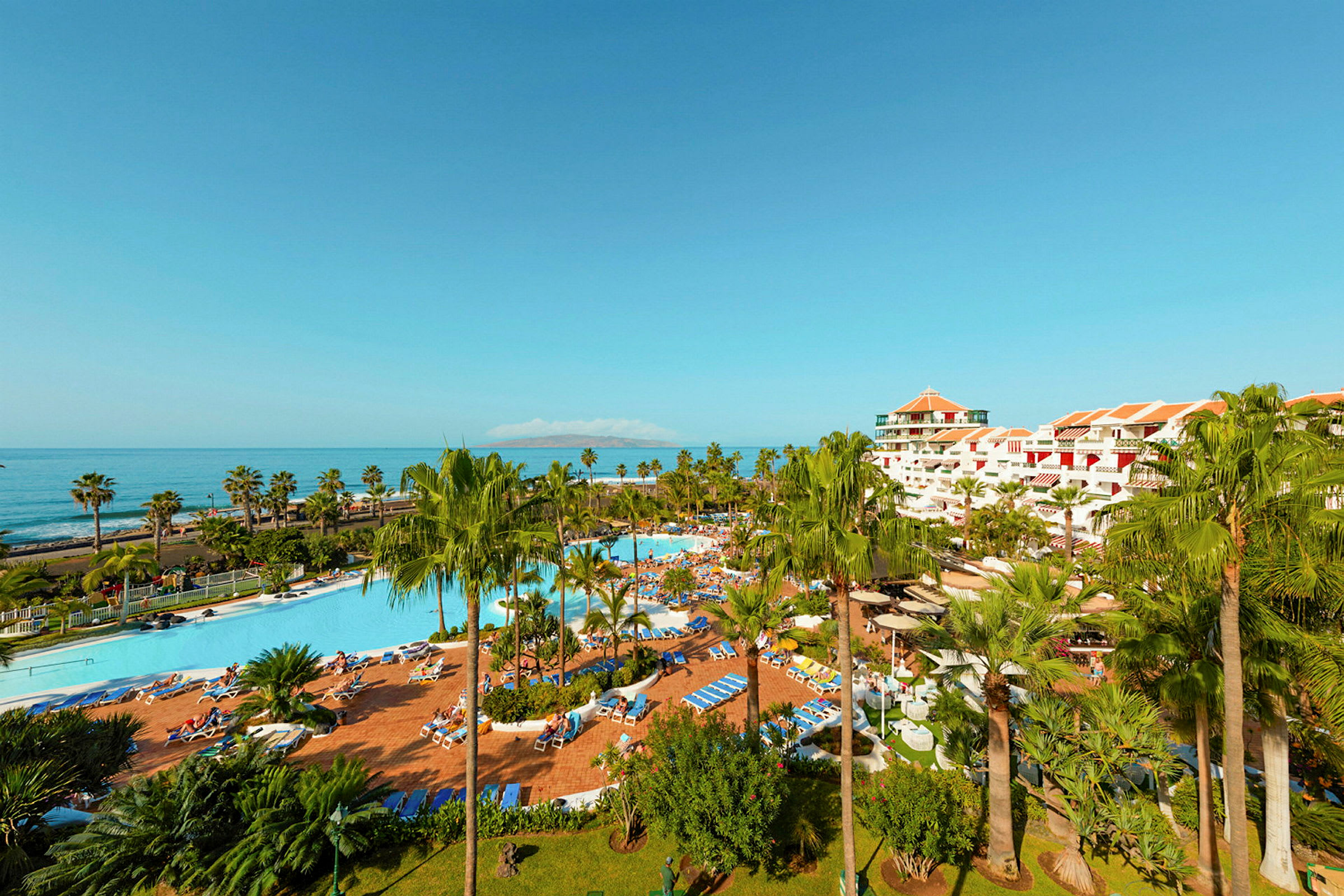 Resort med poolområde, palmer og havet i baggrunden. (AI-genereret tekst)