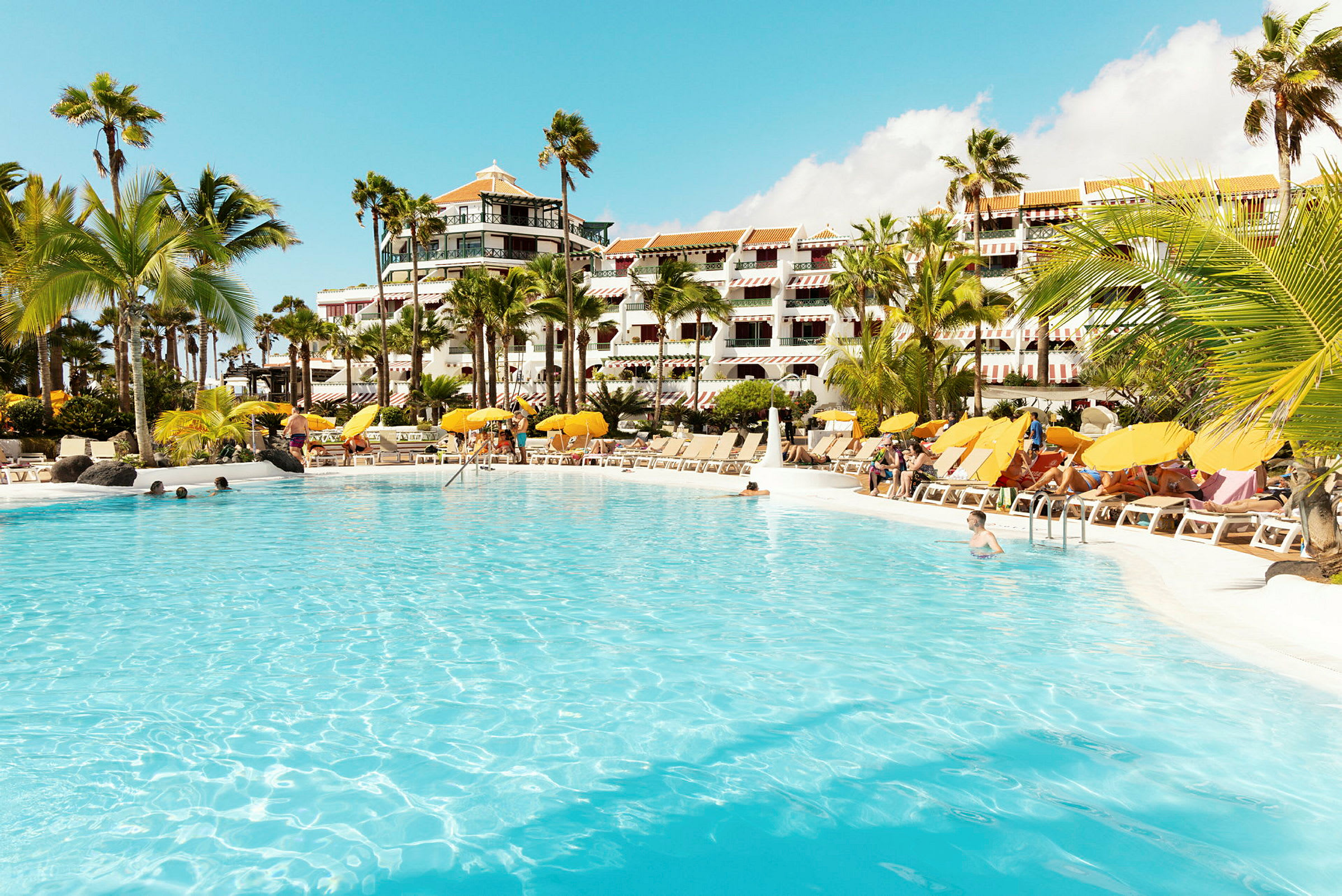 Stor swimmingpool omgivet af liggestole og parasoller foran hotelbygning. (AI-genereret tekst)