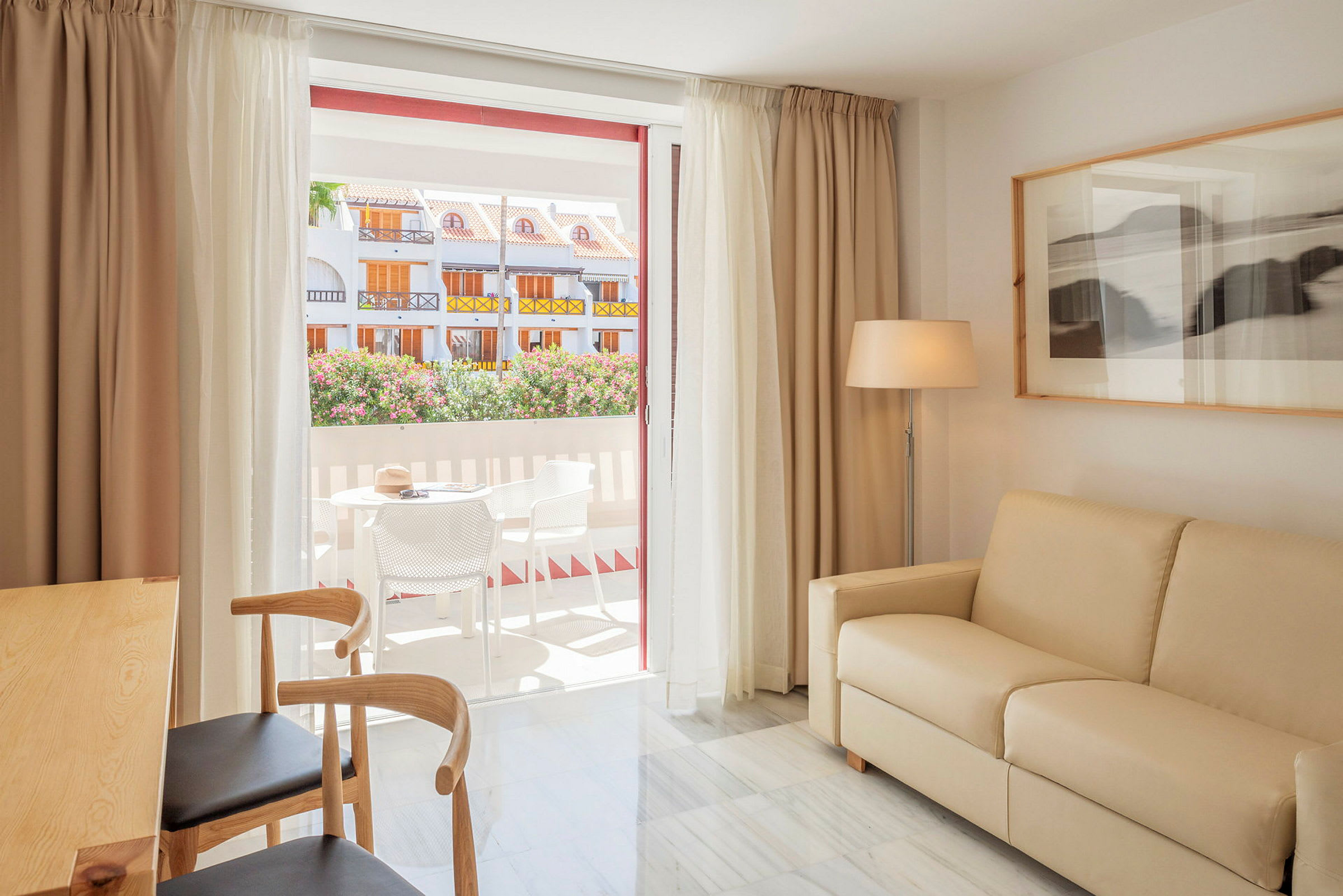 Et rummeligt og lyst opholdsområde med beige sofa, træstole og et lille spisebord. Store glasdøre fører ud til en terrasse med havemøbler, hvor man kan se blomstrende buske og farverige bygninger. På væggen er der et indrammet billede og en gulvlampe. (AI-genereret tekst)