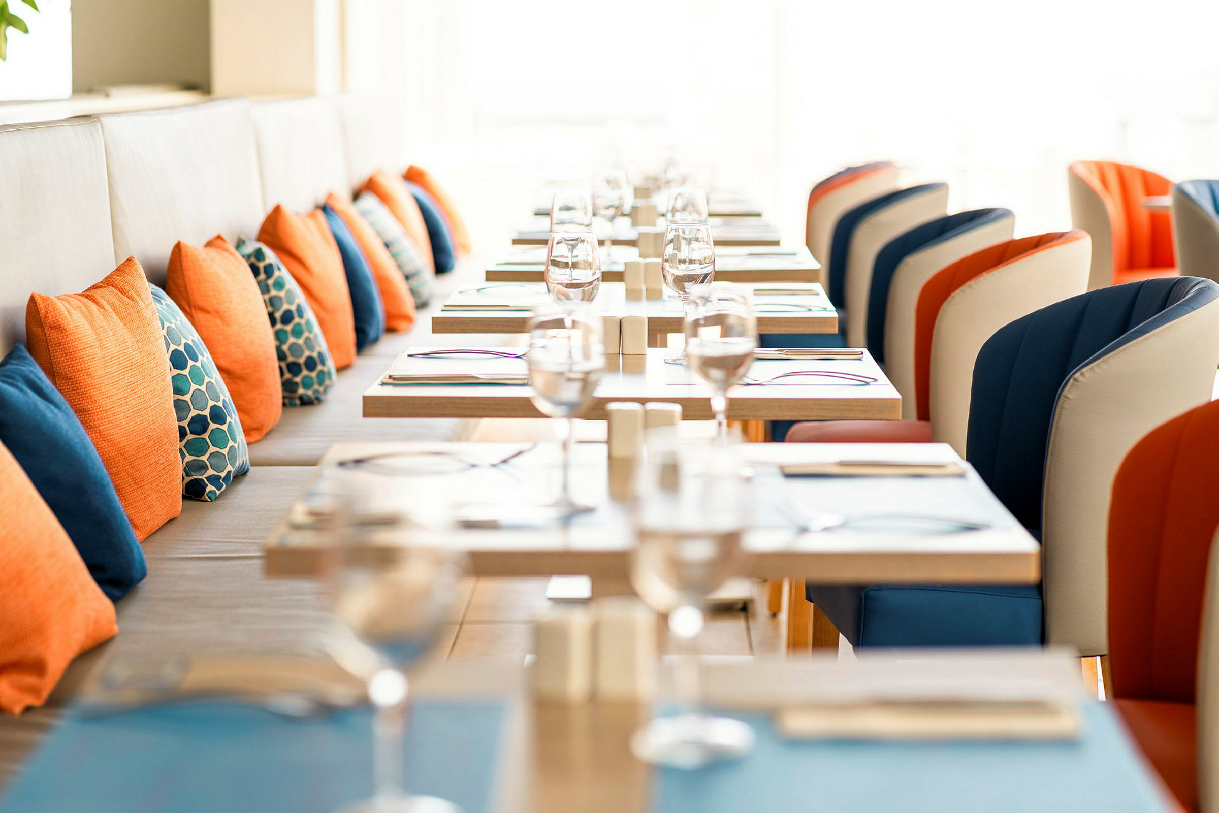 Billedet viser en opstillet restaurant med borde og stole. Stolene er i orange og blå farver, og orange og blå puder pryder siddepladserne ved væggen. (AI-genereret tekst)