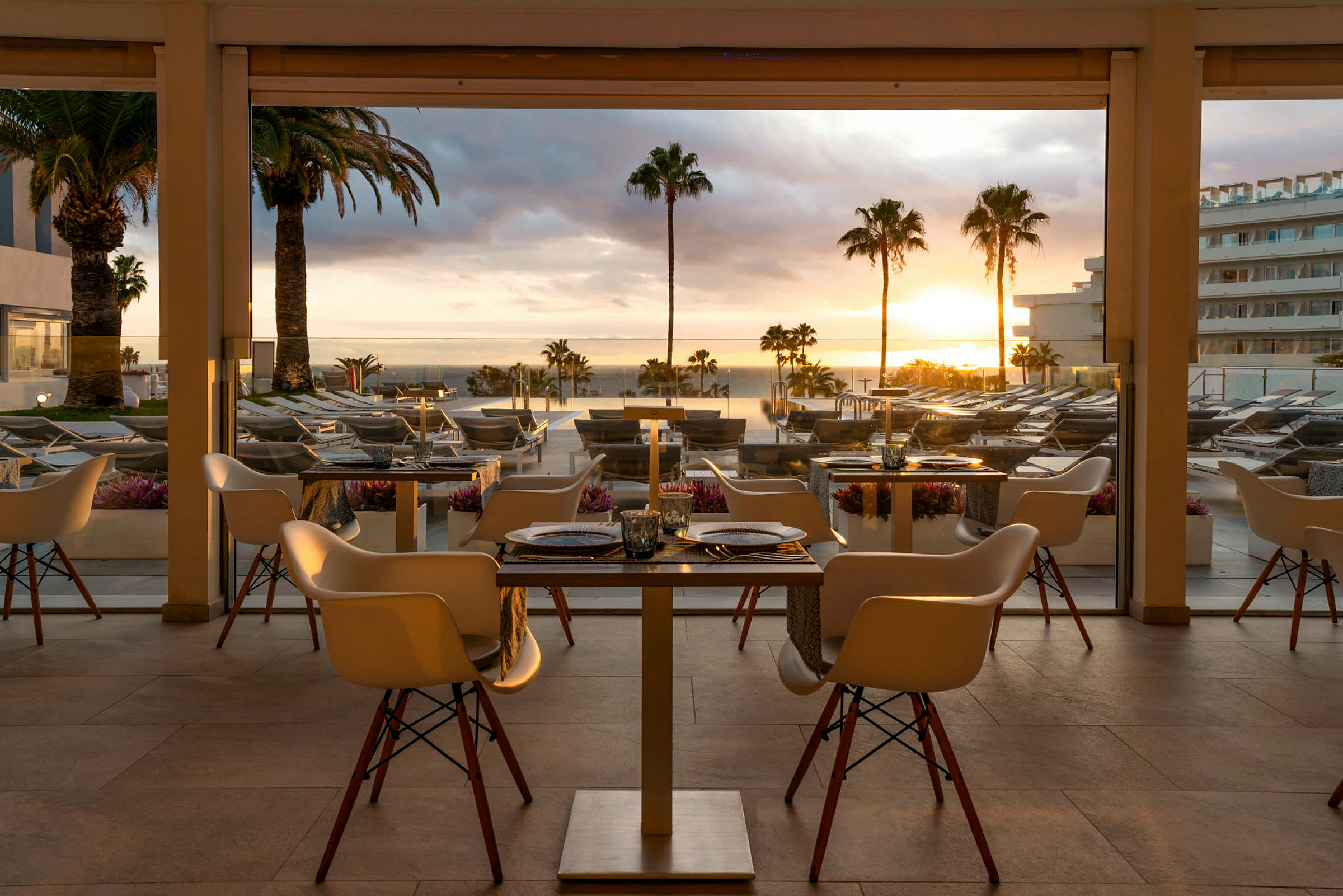 Billede af en restaurant med borde og stole, ved solnedgang med udsigt over en terrasse og en pool omgivet af palmer. (AI-genereret tekst)