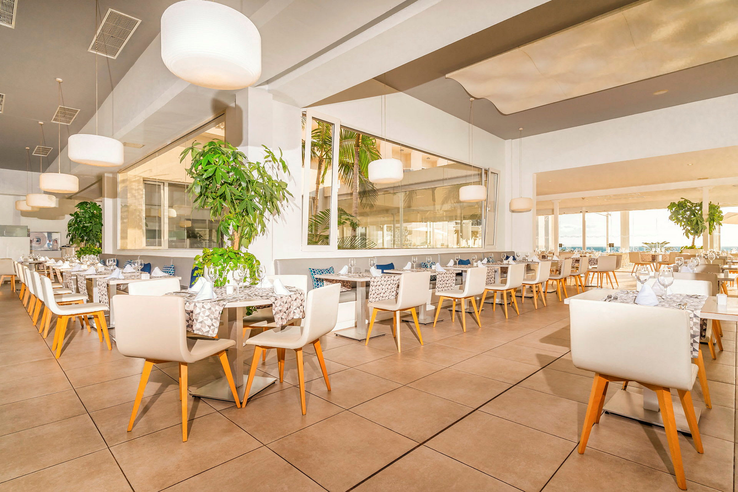 En lys og stilfuld restaurant med hvide borde og stole, omgivet af grønne planter. (AI-genereret tekst)