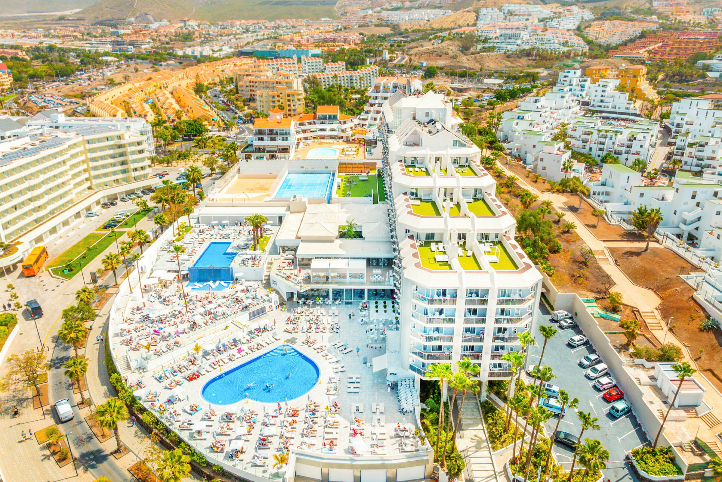 Et luftfoto af et hotelområde med pools, liggestole og omkringliggende bygninger. (AI-genereret tekst)
