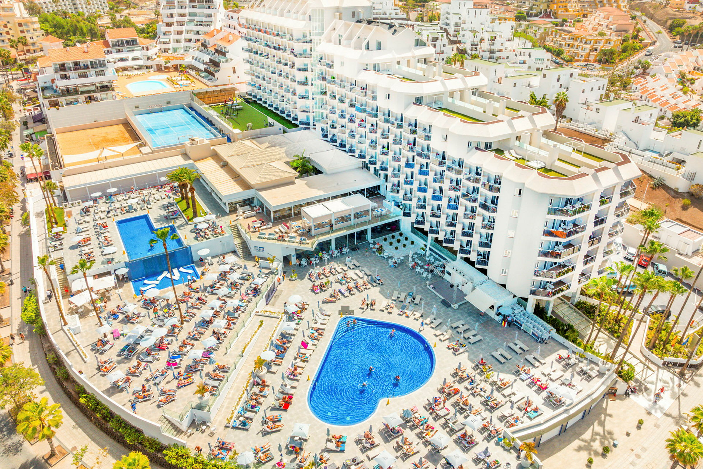 Hotelkompleks med poolområde og liggestole, omgivet af bygninger og grønne områder. (AI-genereret tekst)
