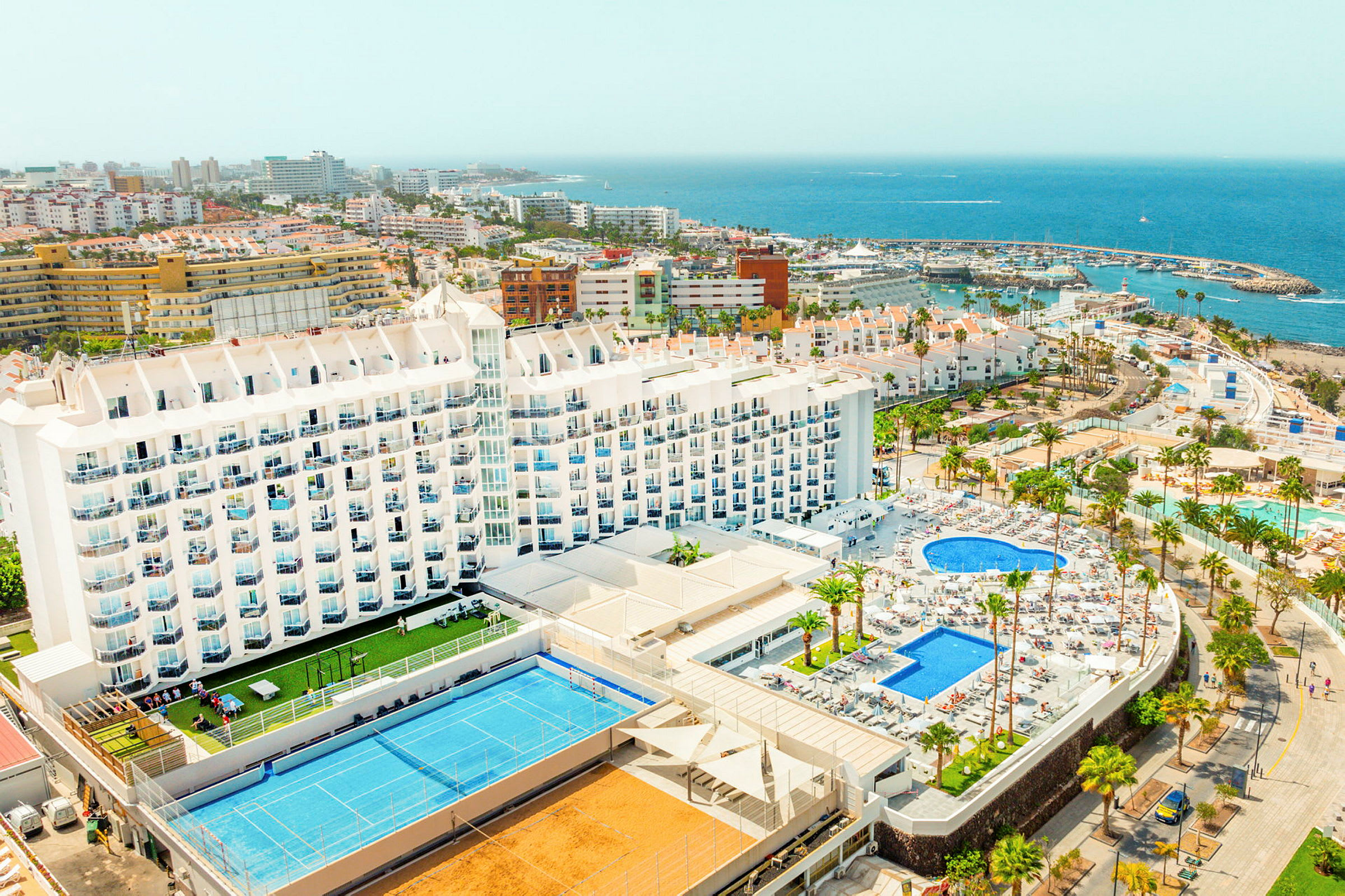 Billede af et stort hotelkompleks med pools og sportsfaciliteter, omgivet af bygninger, palmer og kystlinje. (AI-genereret tekst)