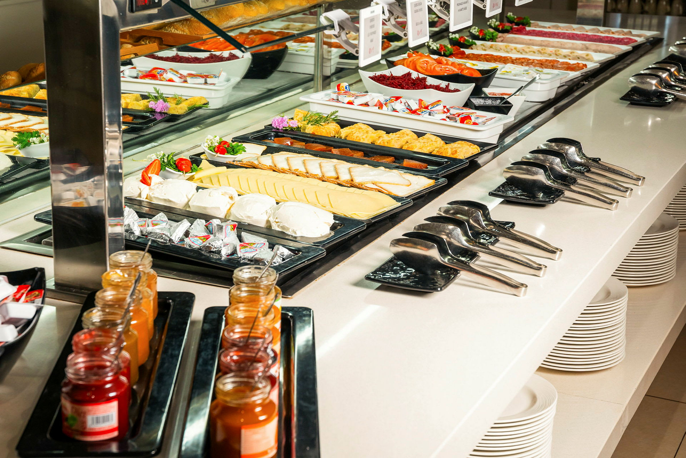 En buffet med forskellige retter, herunder skiver af ost, grøntsager og glas med marmelade, med serveringsredskaber og stablede tallerkener.  (AI-genereret tekst)