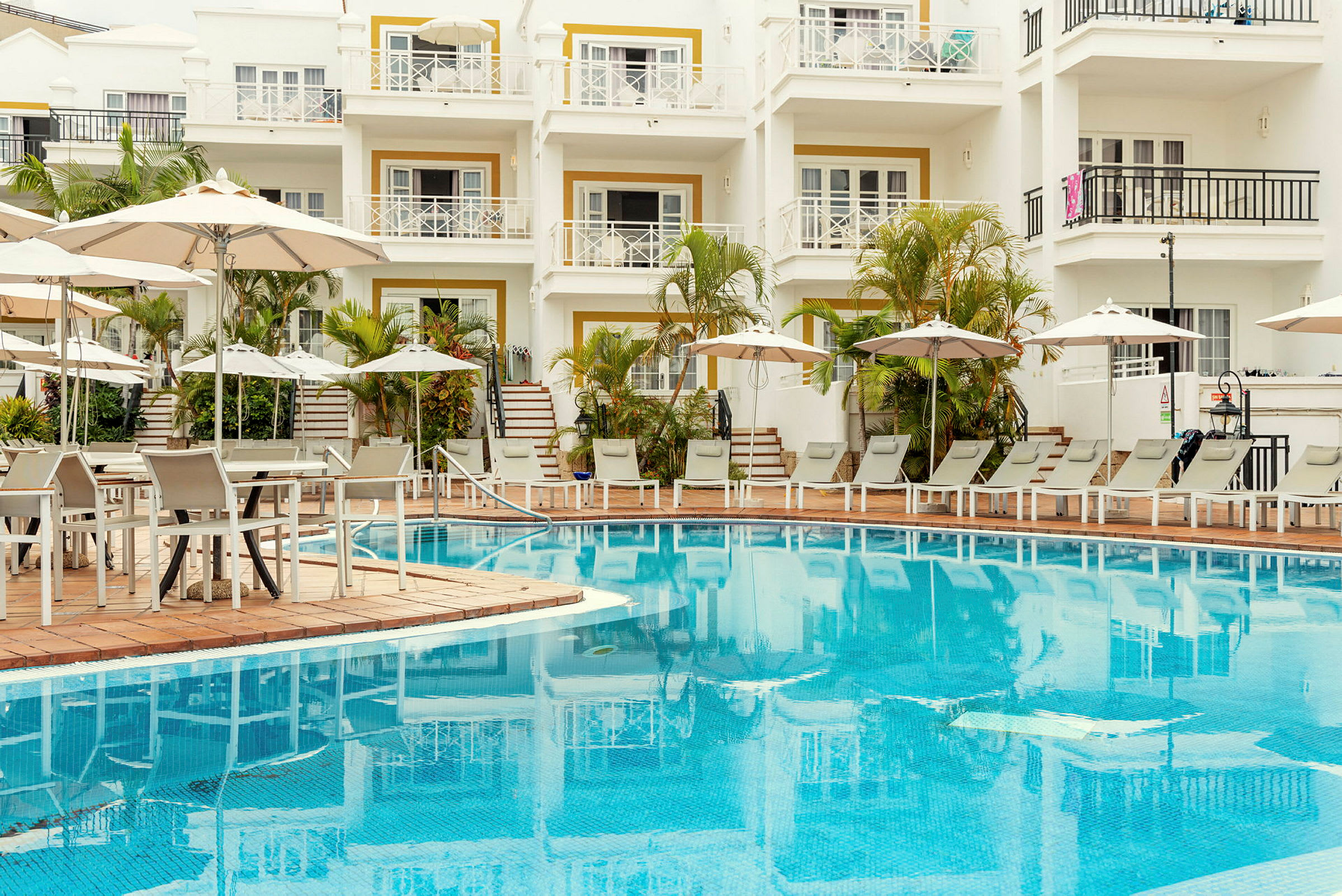 Billede af en hotelpool med liggestole, parasoller og en boligblok i baggrunden. (AI-genereret tekst)