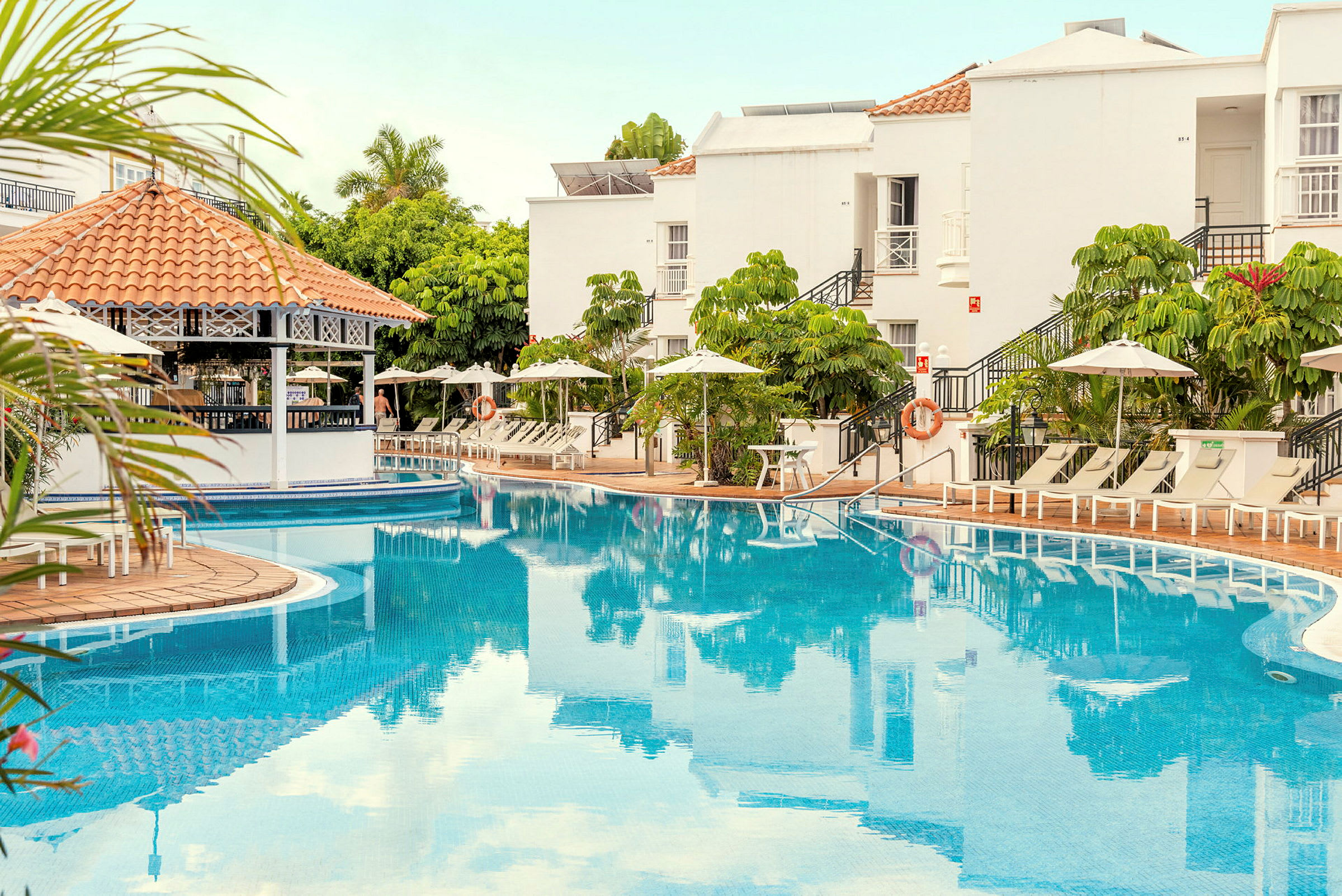 Udendørs swimmingpool med liggestole og parasoller samt hvid bygning i baggrunden. (AI-genereret tekst)