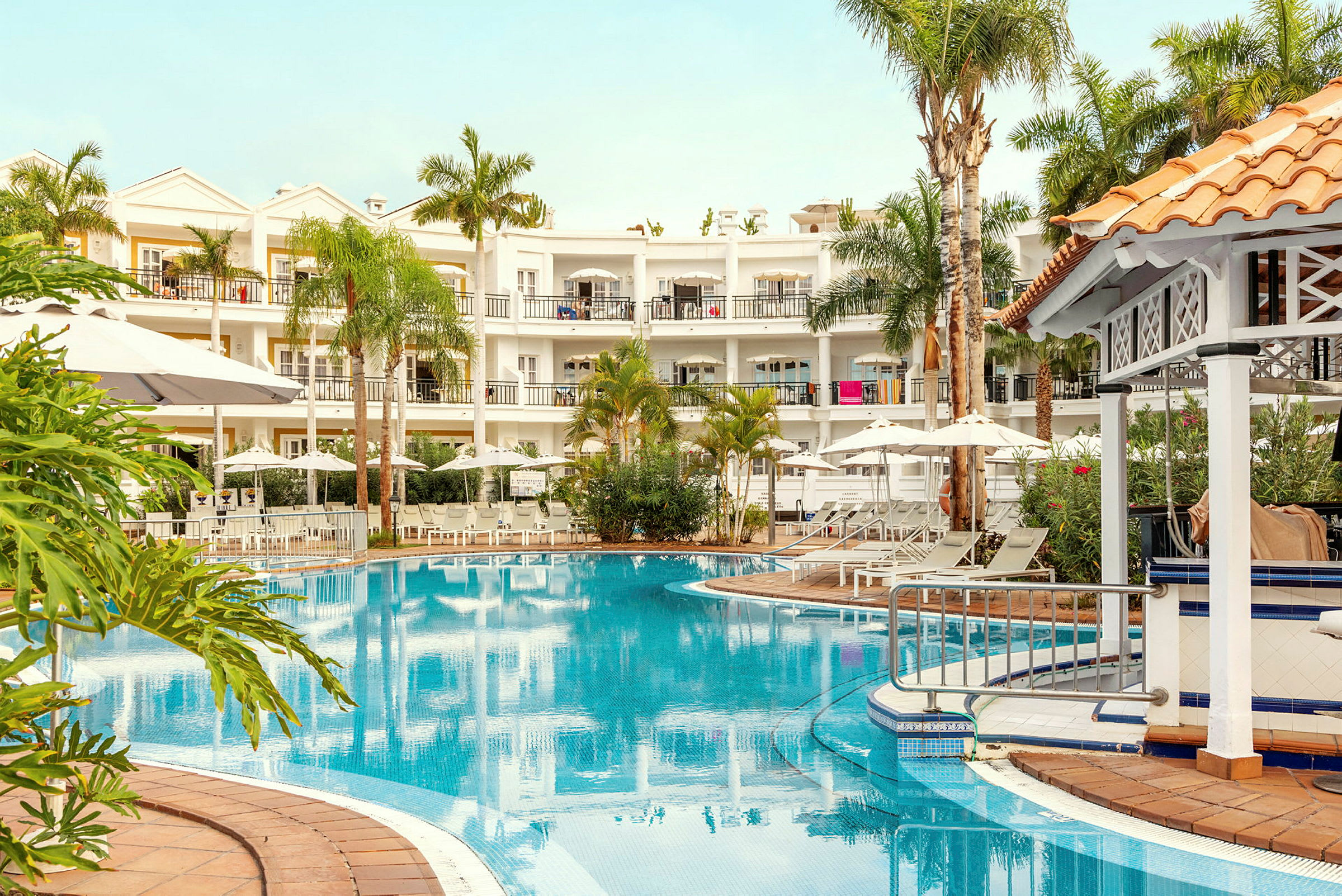 Billede af et hotel med en pool, omgivet af liggestole, parasoller og beplantning. (AI-genereret tekst)