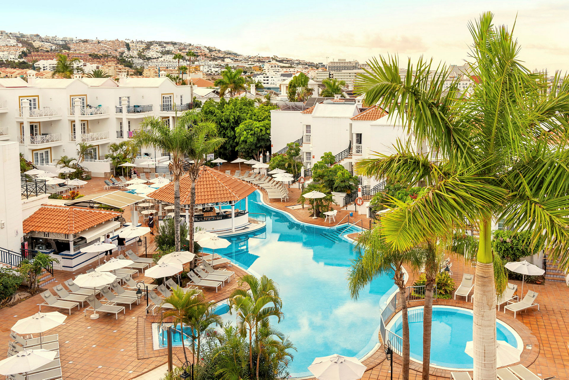 Billede af hotelområde med pool, liggestole og omkringliggende palmer. (AI-genereret tekst)